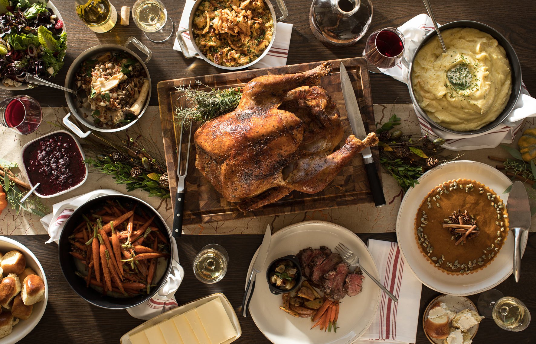 A table filled with various dishes including a roasted turkey, green salad, stuffing, mashed potatoes, carrots, cranberry sauce, and apple pie, with glasses of wine. The table is decorated with greenery and napkins.