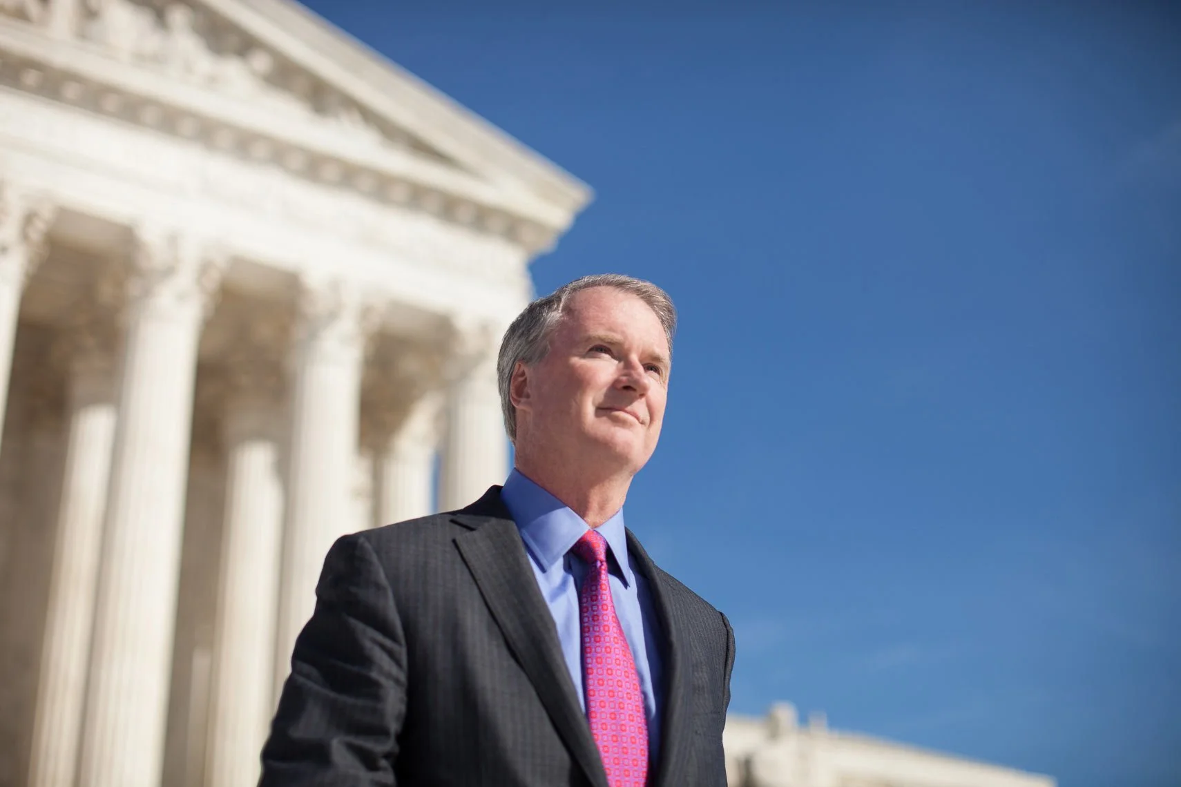 A suited man with gray hair wearing a blue shirt and red tie standing outside in front of the U.S. Capitol building, looking into the distance against a clear blue sky.