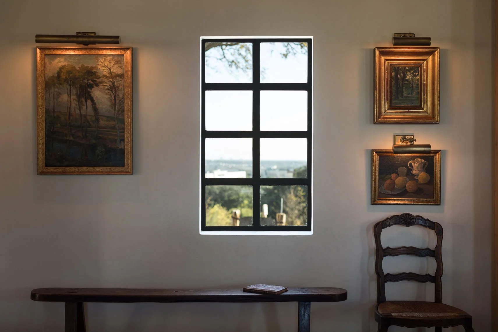 Interior wall with four framed landscape paintings (two large and two small) illuminated by picture lights, a window in the center showing an outdoor view of trees and buildings, and a wooden chair with a woven seat to the right of the paintings.