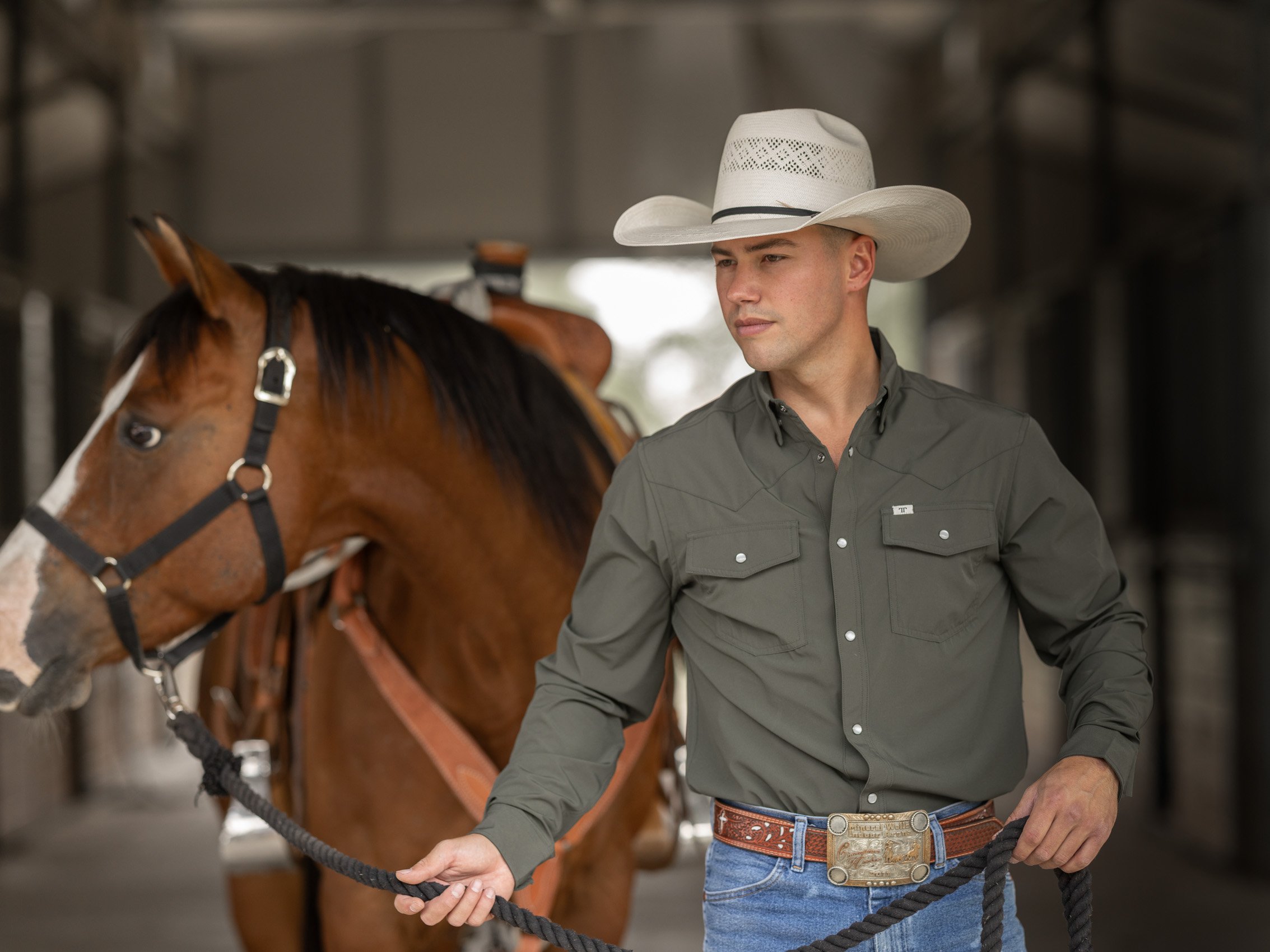 A man in a gray western shirt, wearing a large white cowboy hat with decorative cutouts, holding a black rope, standing next to a brown horse with a black mane and bridle inside a stable.