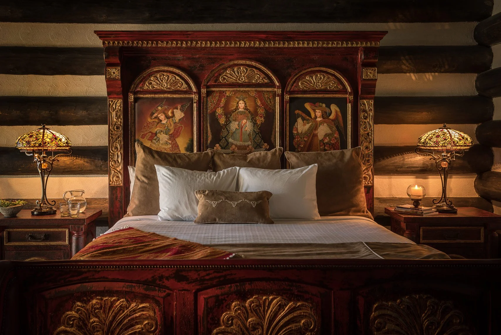 A luxurious bed with a wooden headboard adorned with three religious icons, flanked by two ornate table lamps, and decorated with many pillows, in a cozy log cabin-style bedroom.