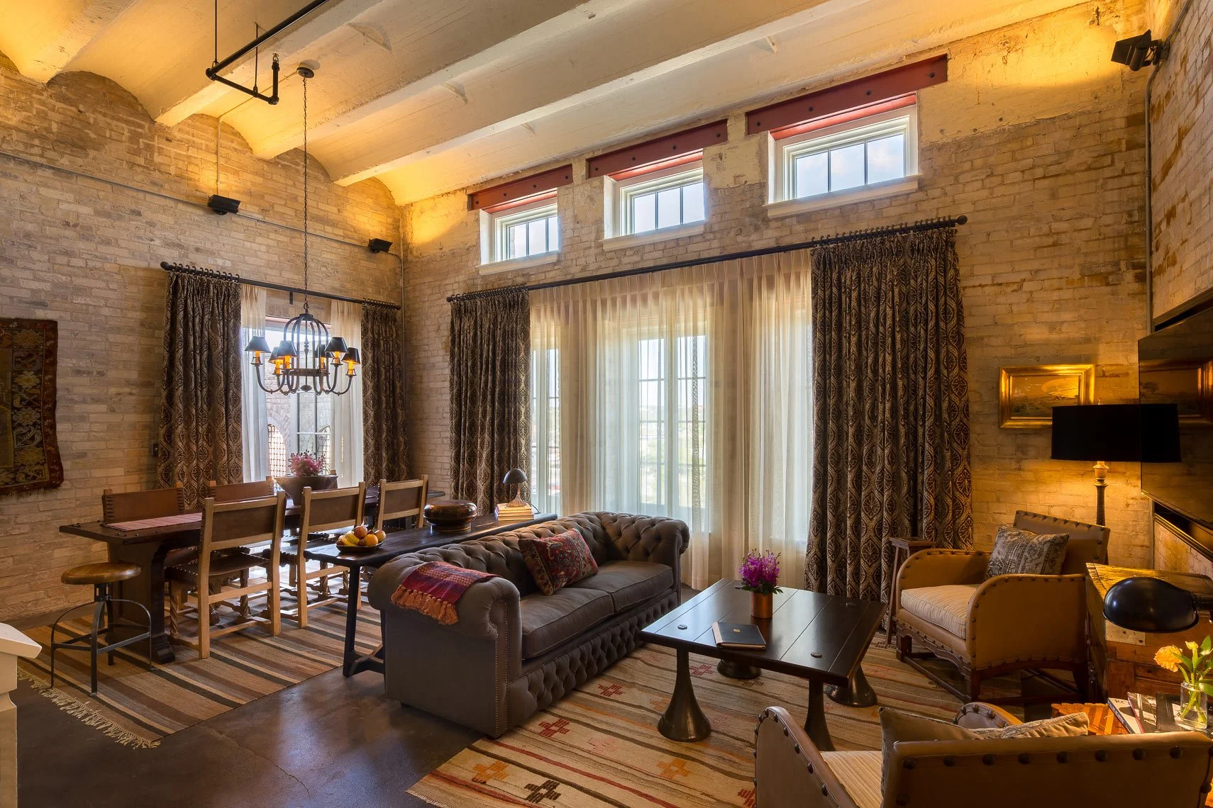 A cozy living and dining room with exposed brick walls, vaulted ceiling, large windows with curtains, a chandelier, a gray tufted sofa with throw pillows, a wooden coffee table with a flowerpot, separate armchairs, a dining table with wooden chairs, 