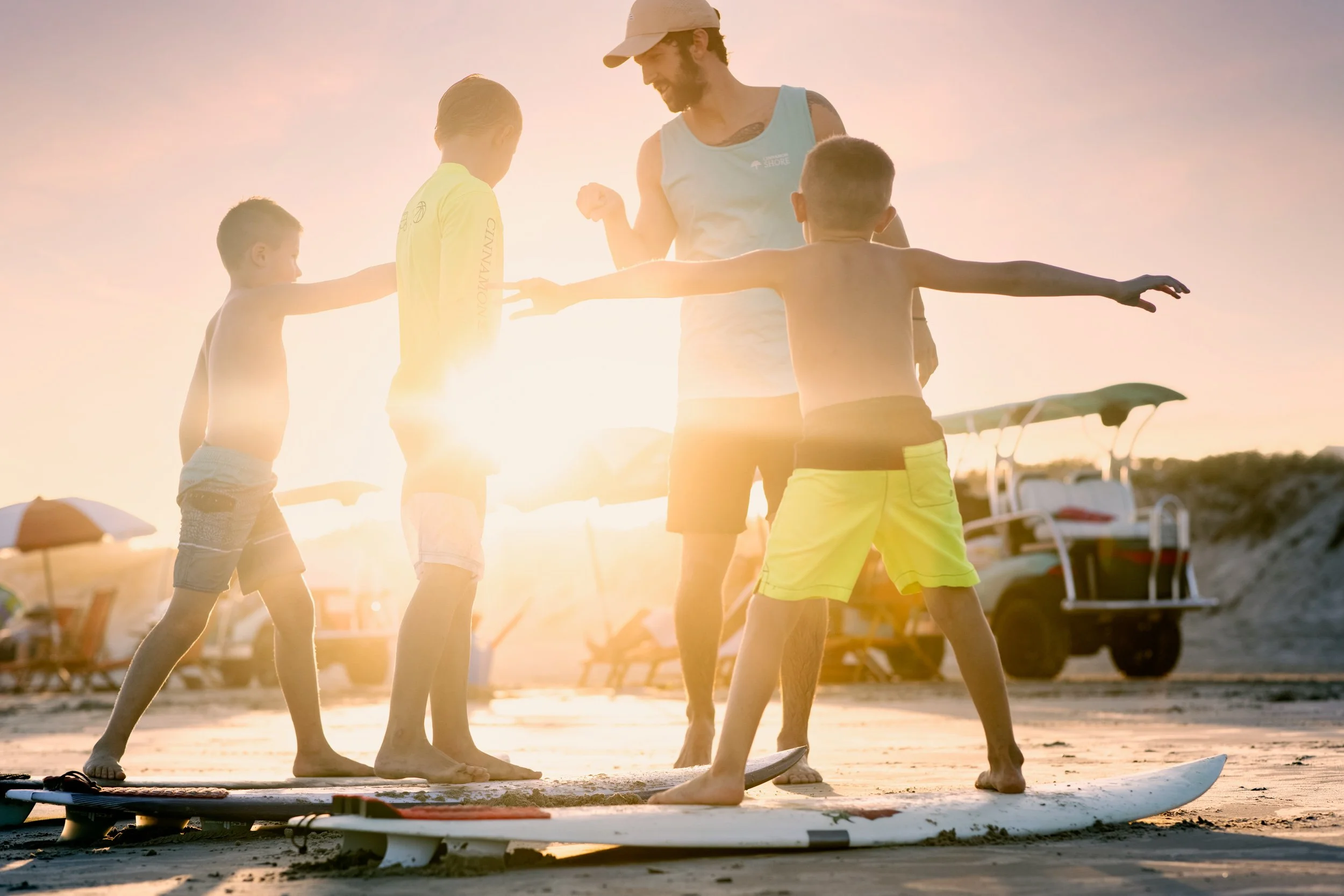 A group of children and a man with a beard, possibly a surf instructor, gathered on a beach at sunset, practicing surfing. The kids are shirtless, wearing shorts, and standing on surfboards, while the man and some children wear tank tops and hats. Th