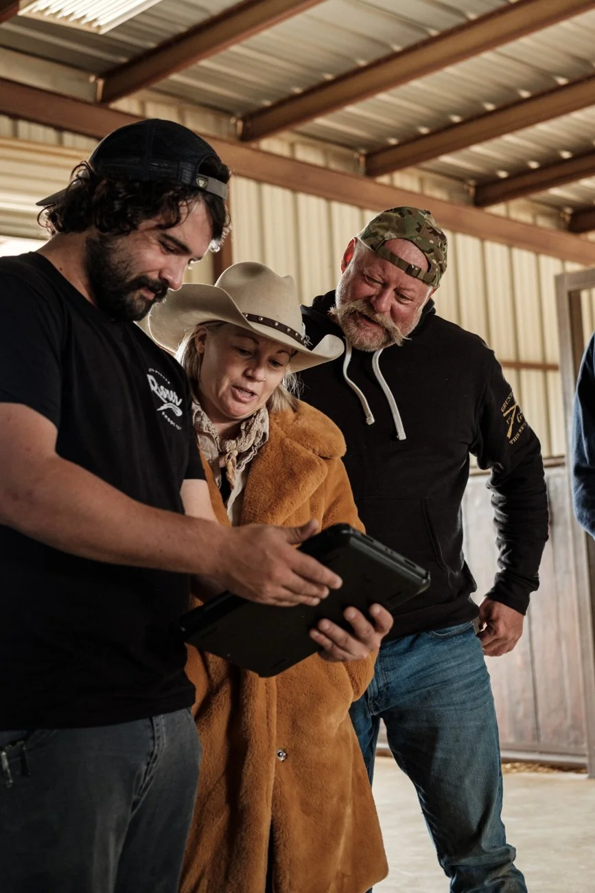 Three people looking at a device inside a barn, two men and a woman, with the woman wearing a cowboy hat and a brown coat.
