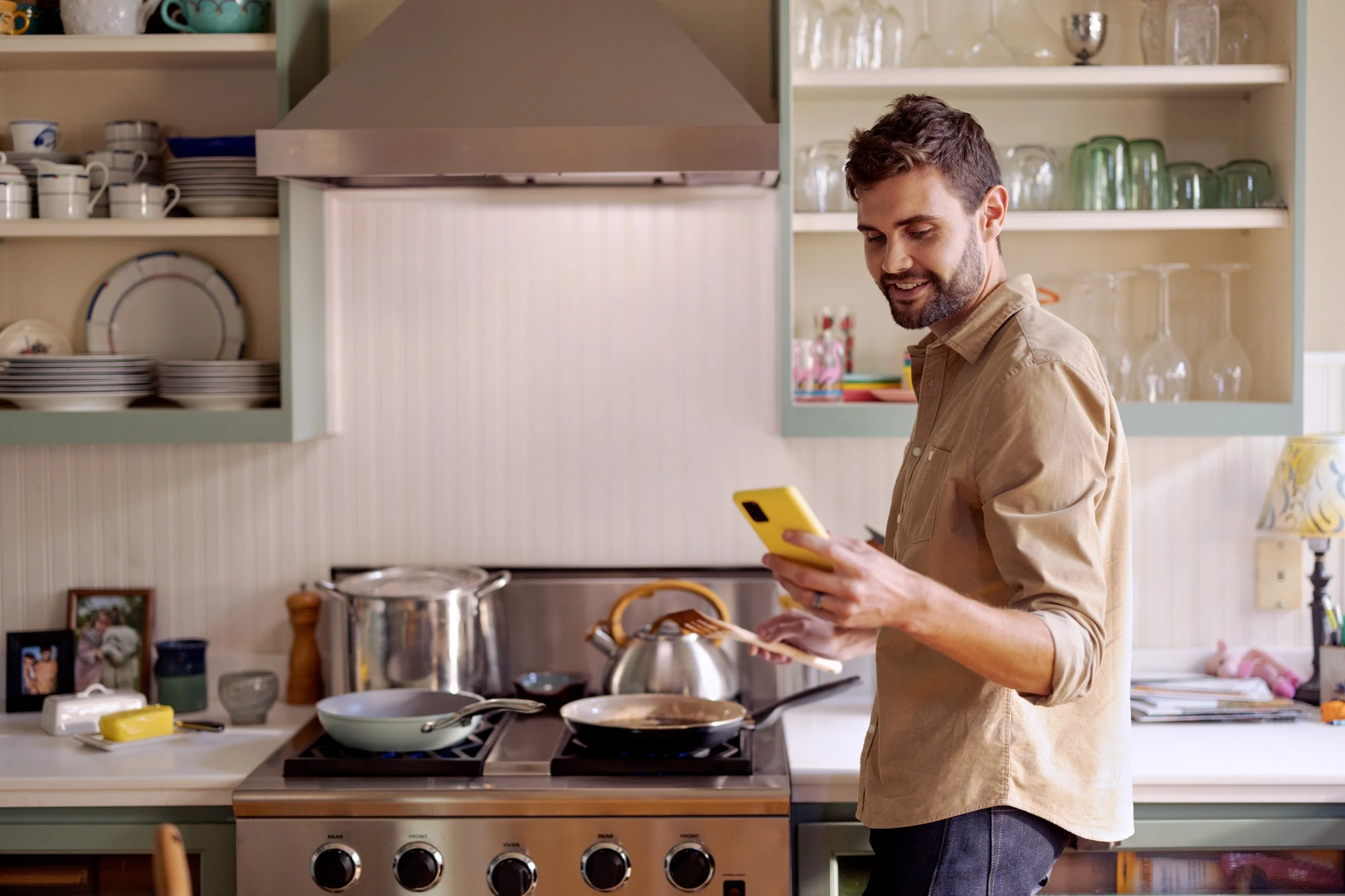 A man with brown hair and a beard, wearing a beige shirt, is standing in a kitchen, looking at his yellow phone. The kitchen has open shelves with dishes, glasses, and decorative items.