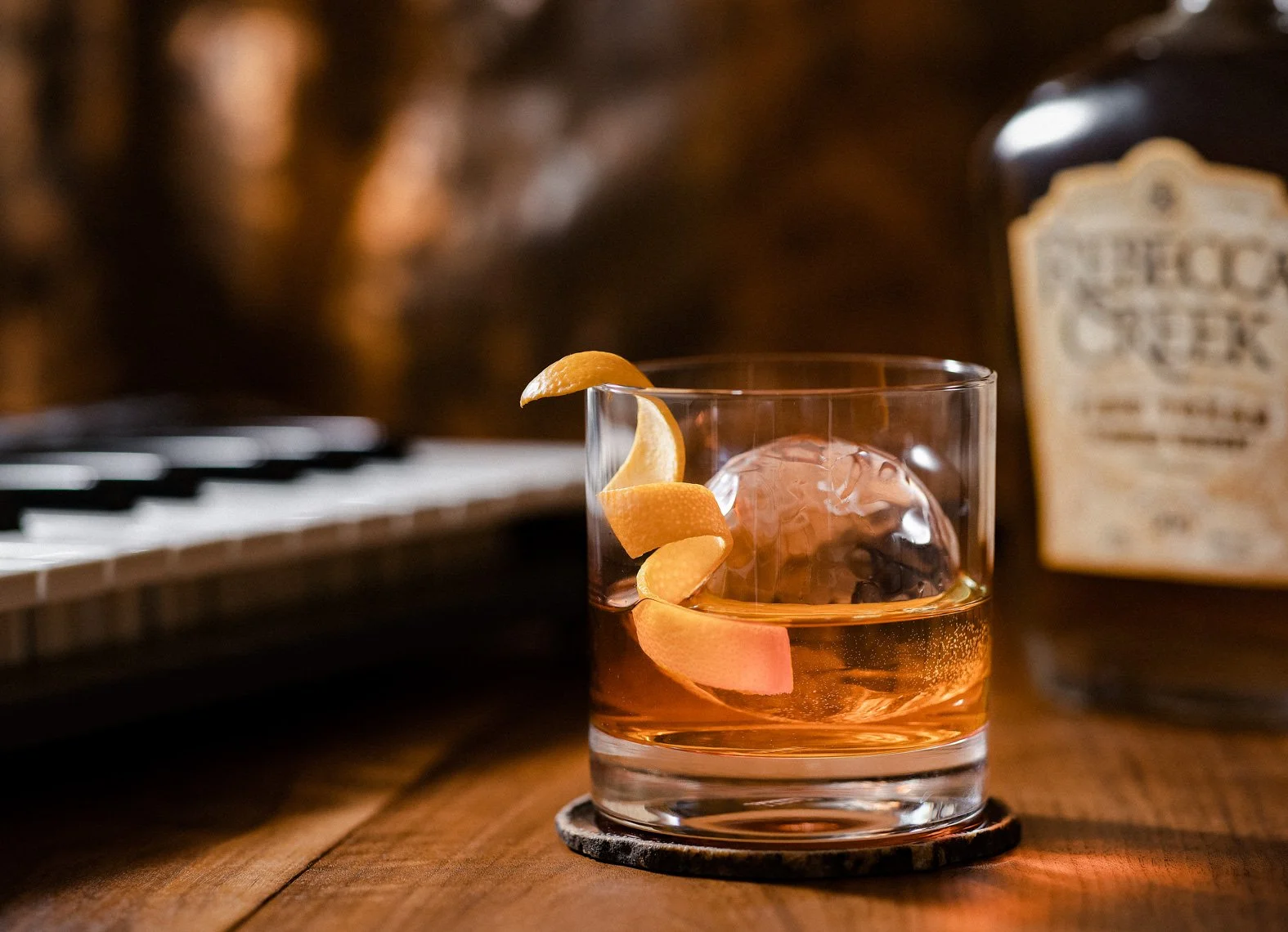 A glass of whiskey with a large ice cube and orange peel garnish on a coaster, with a bottle of whiskey in the background and a keyboard on the left side.