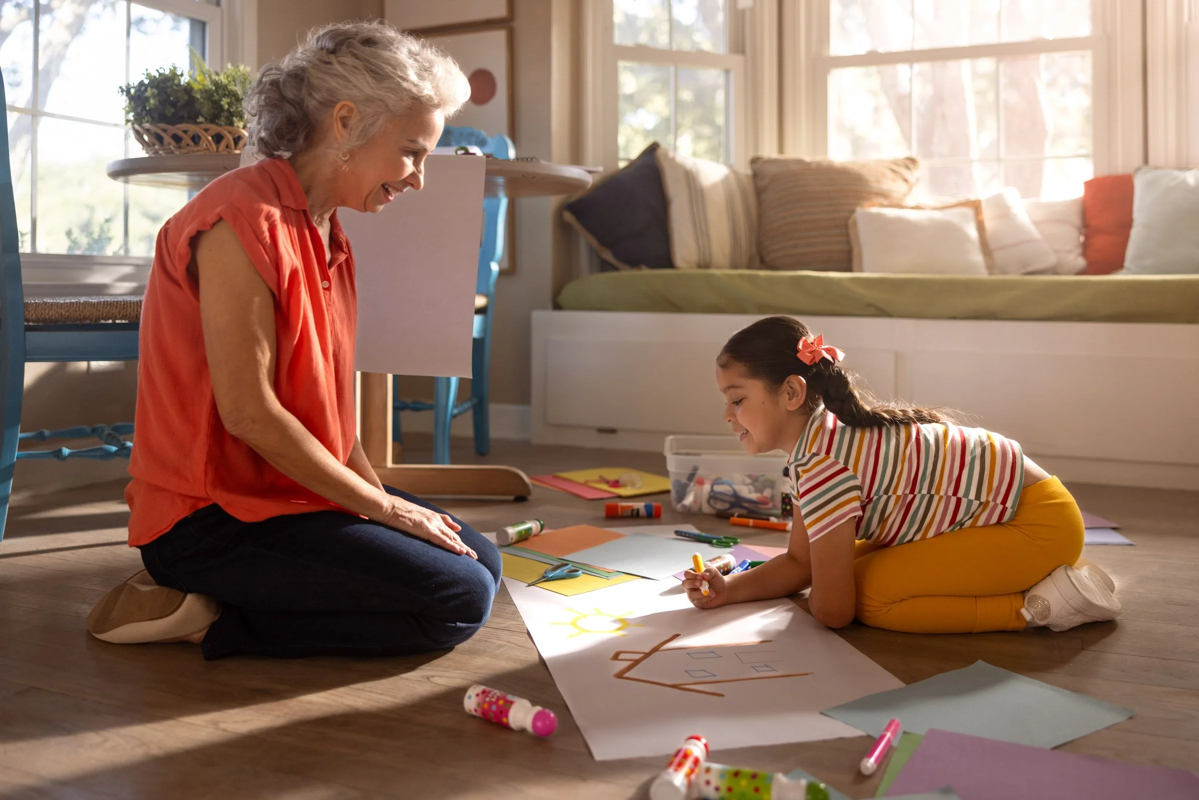 An elderly woman and a young girl are sitting on the floor in a sunlit room, drawing and coloring with various art supplies on a large sheet of paper.