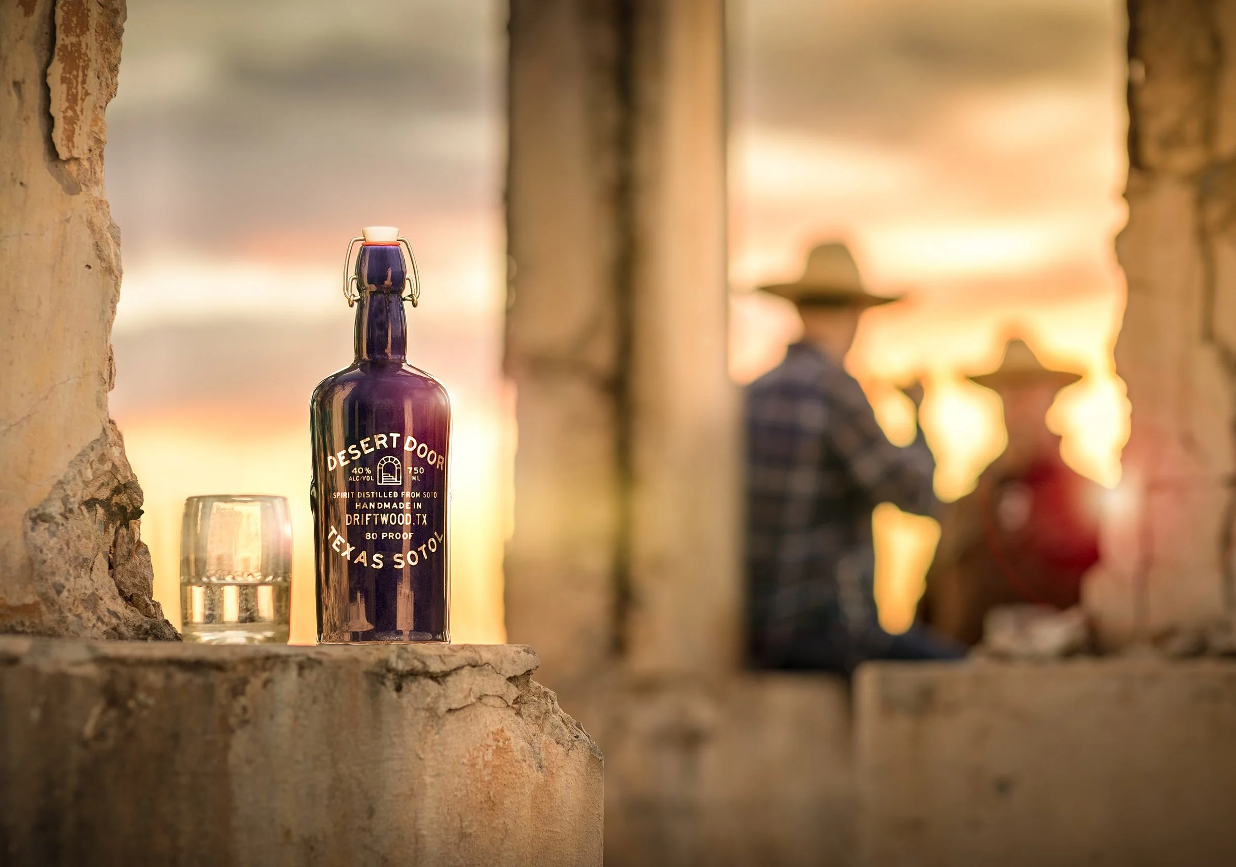 Close-up of a purple bottle labeled 'Desert Door' with a glass in front, set against a rustic brick and wood background during sunset.