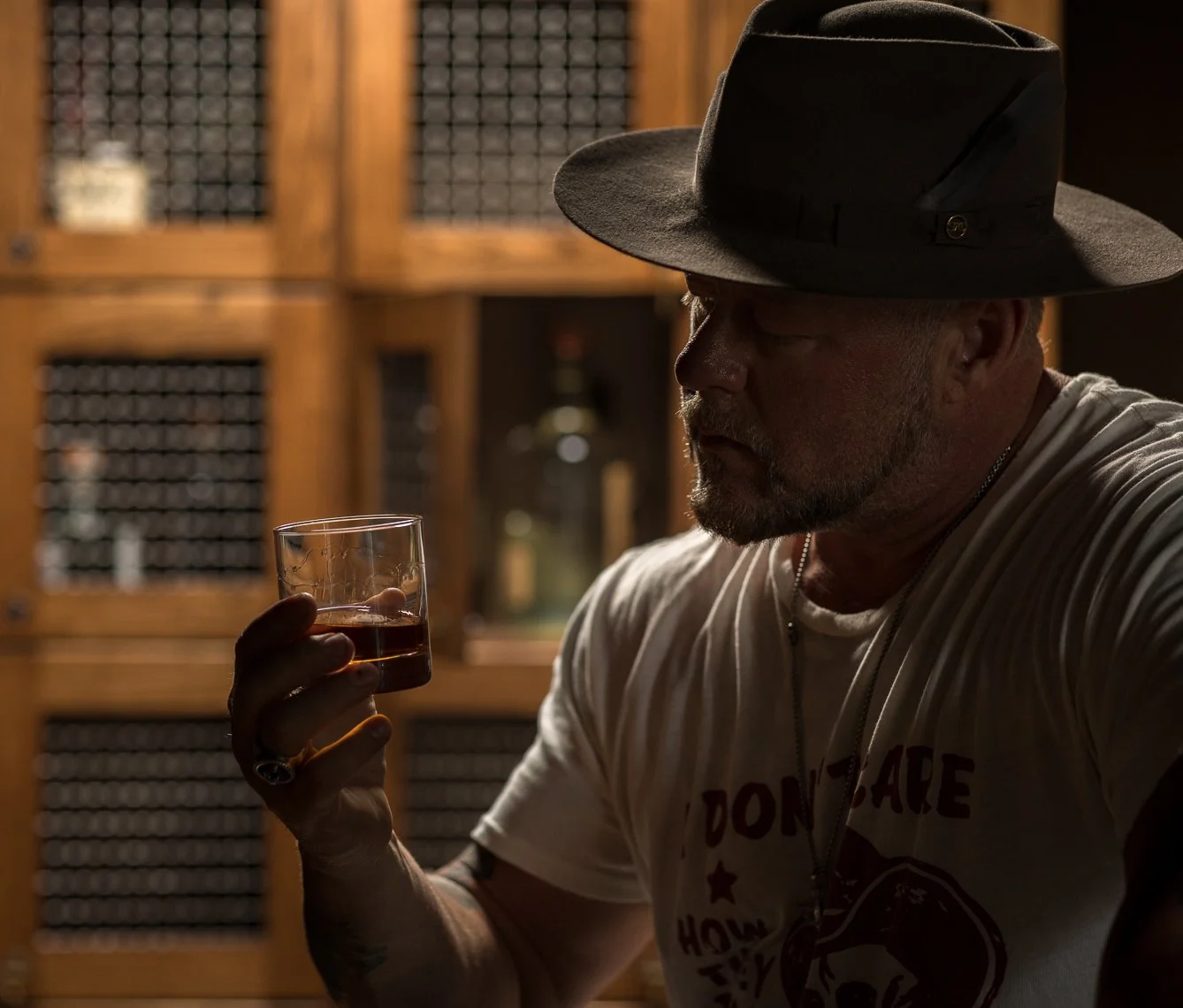 A man with a beard, wearing a wide-brimmed hat and a t-shirt, is holding a glass of whiskey in a dimly lit room with wooden shelves in the background.