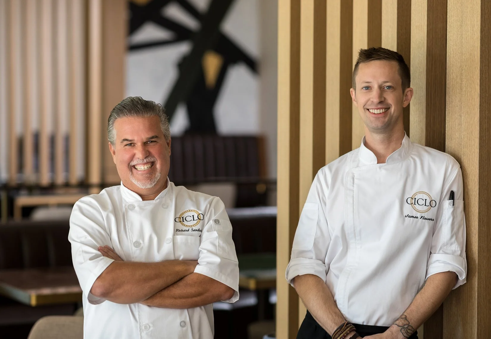 Two male chefs in white uniforms smiling and standing indoors, one with arms crossed, against a wooden slat wall background.
