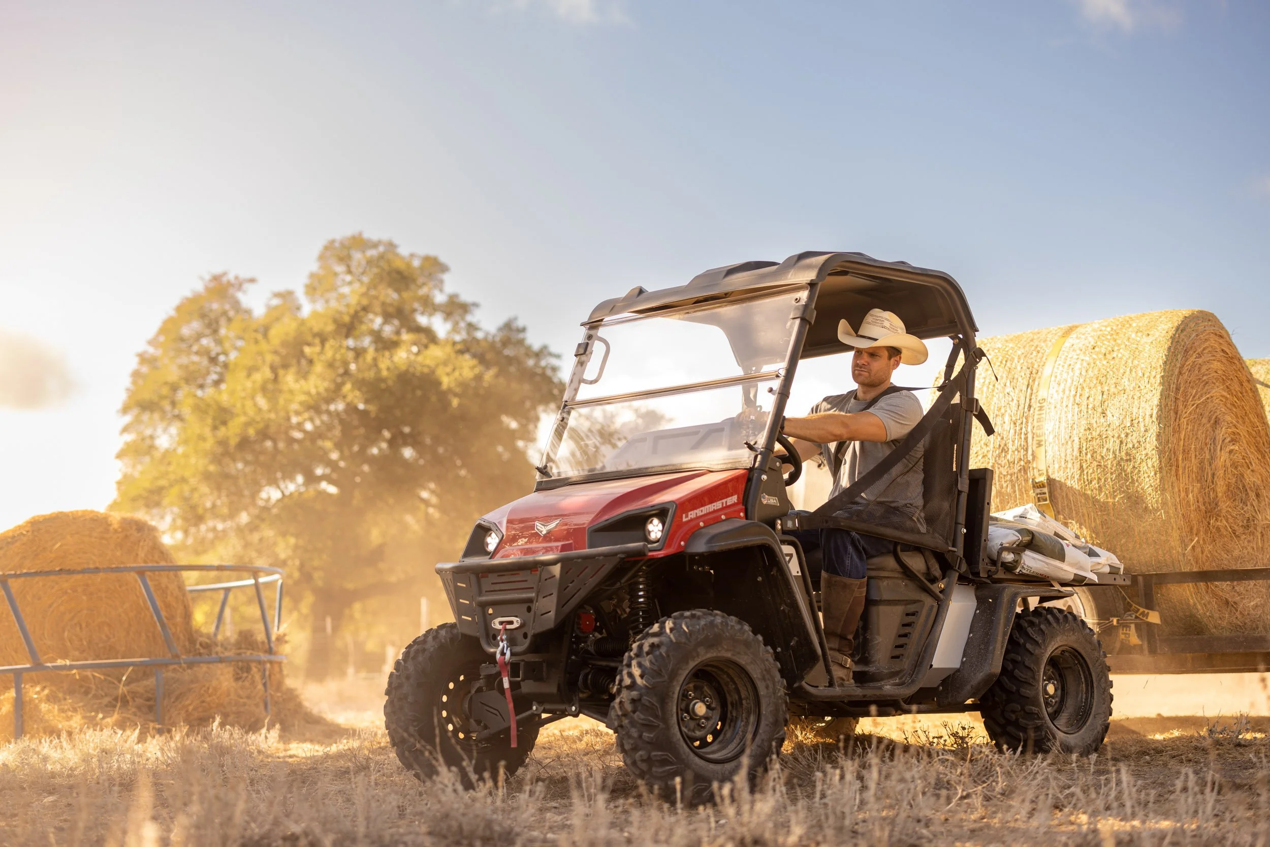 A man wearing a cowboy hat driving a red utility vehicle with large tires, pulling a large hay bale in a farm setting during sunset.