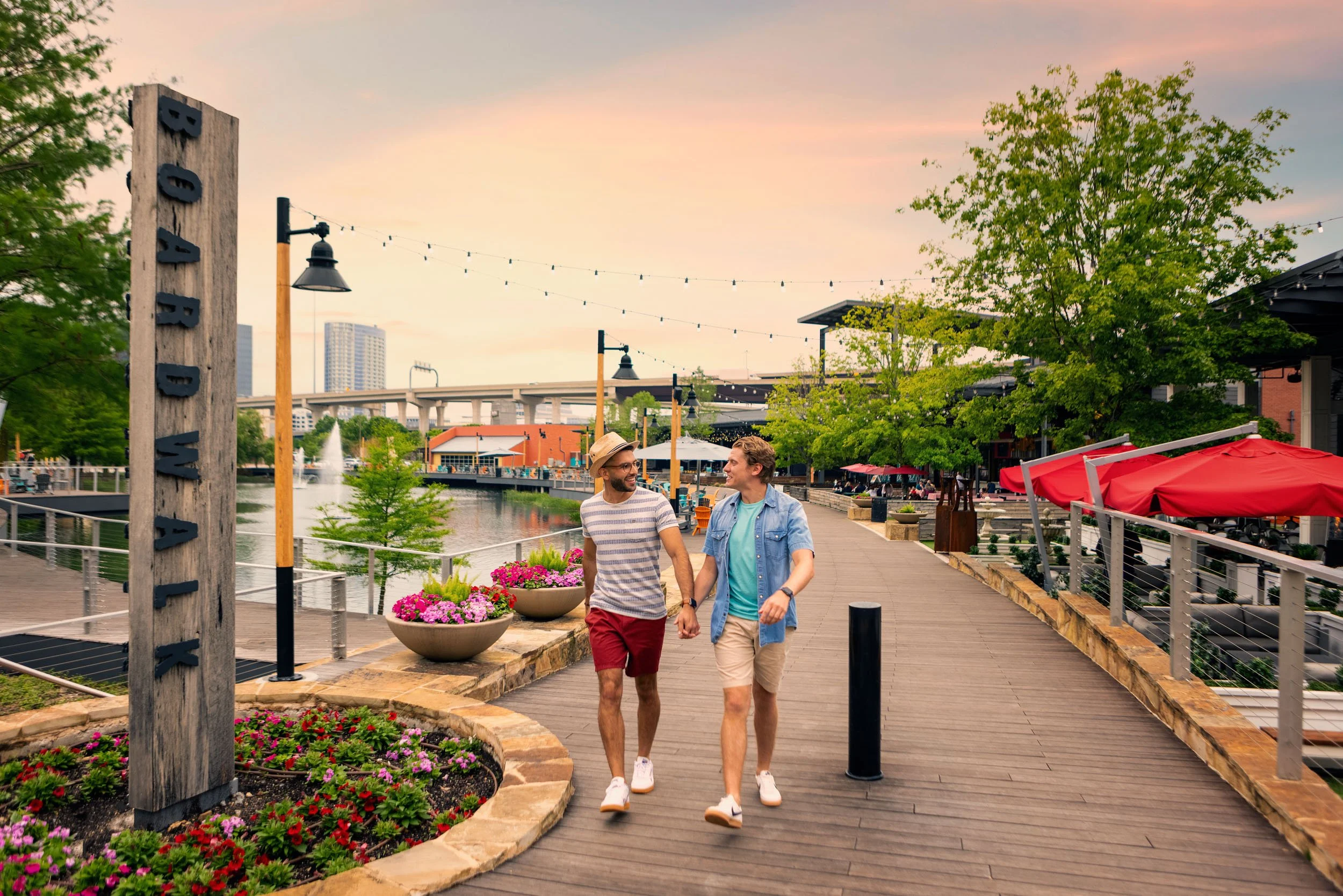 Two young men walking and talking along a waterfront boardwalk at dusk, with greenery, colorful flowers, patio umbrellas, and a city skyline in the background.