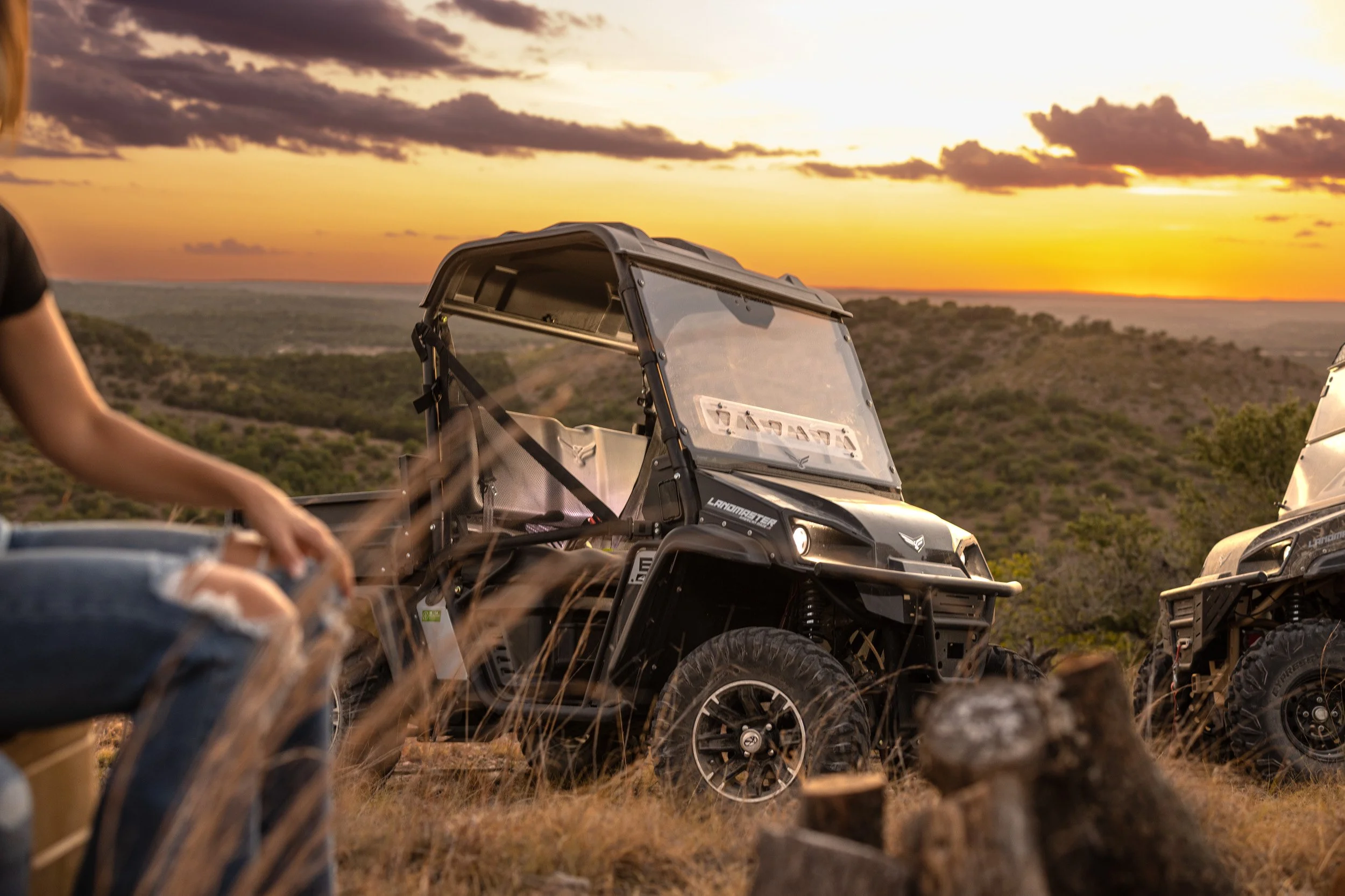 A sunset scene with off-road vehicles parked on a grassy hill, overlooking a scenic landscape with trees and distant hills.