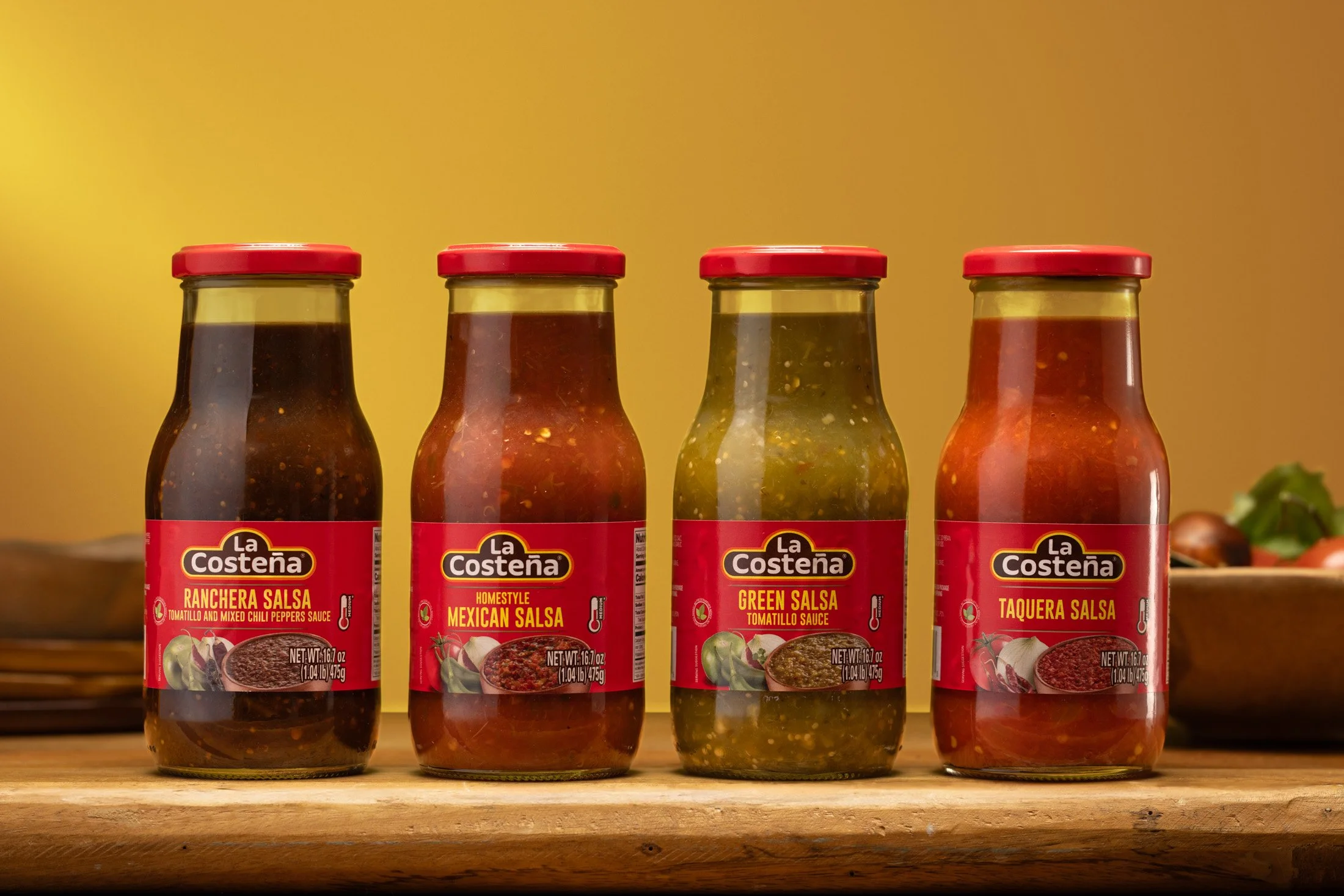 Four jars of La Costeña salsa flavors on a wooden surface against a yellow background. From left to right: Ranchera Salsa, Mexican Salsa, Green Salsa, Taquera Salsa.