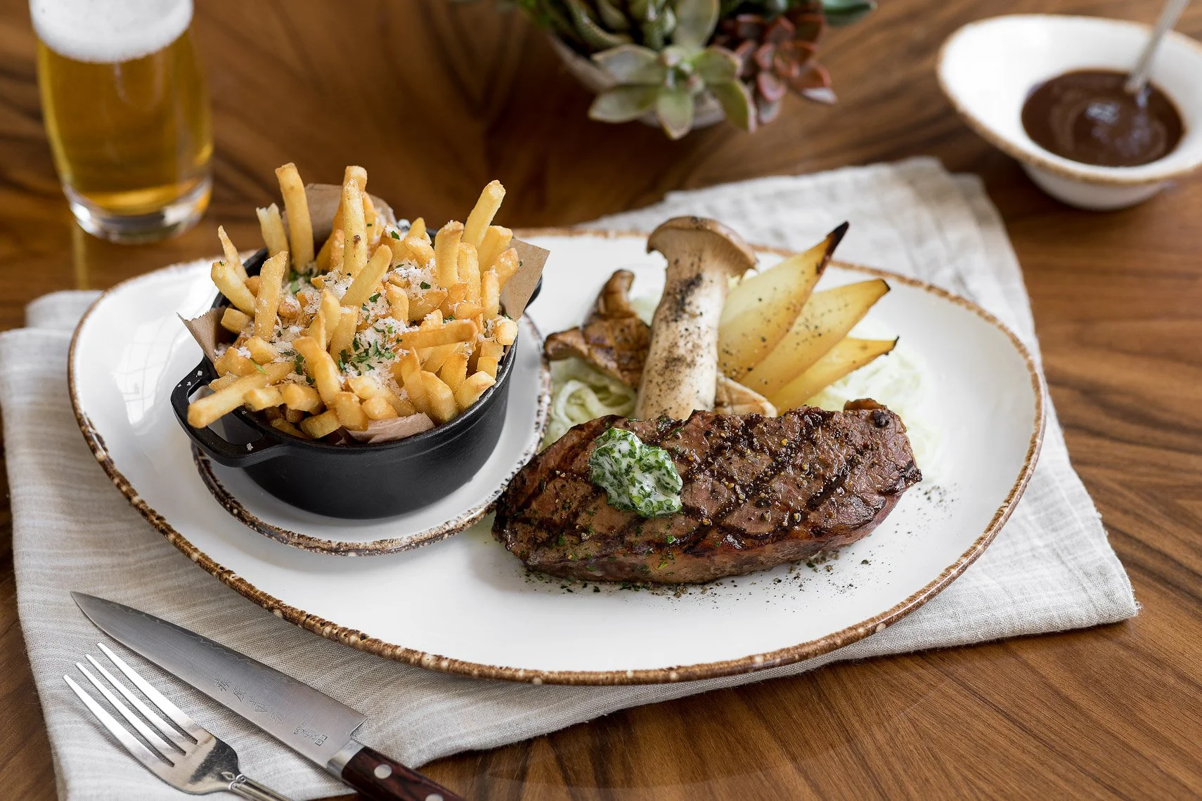 A plate of grilled steak with a dollop of garlic herb butter, served with potato wedges, grilled mushrooms, and a side of fries topped with cheese and herbs. In the background, a glass of beer and a bowl of brown sauce are visible.