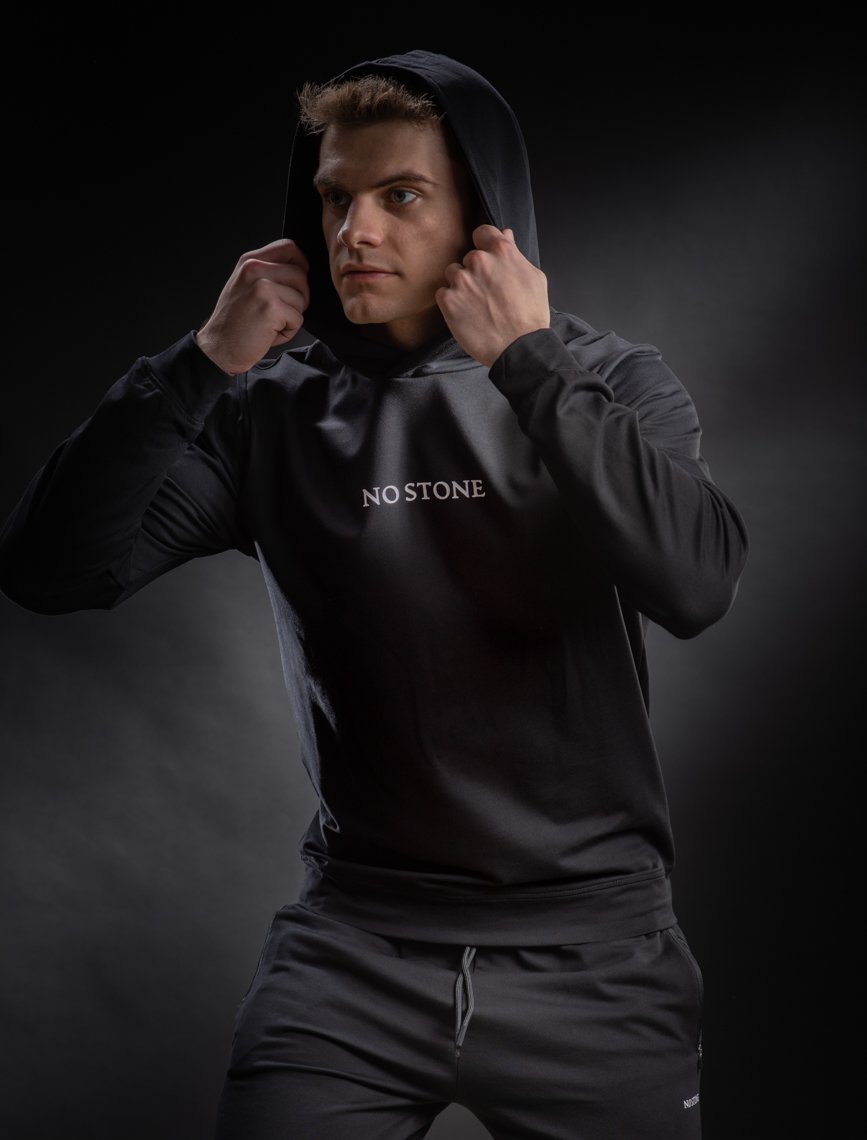 Young man in black hoodie and sweatpants pulling the hood over his head, standing against a dark background.