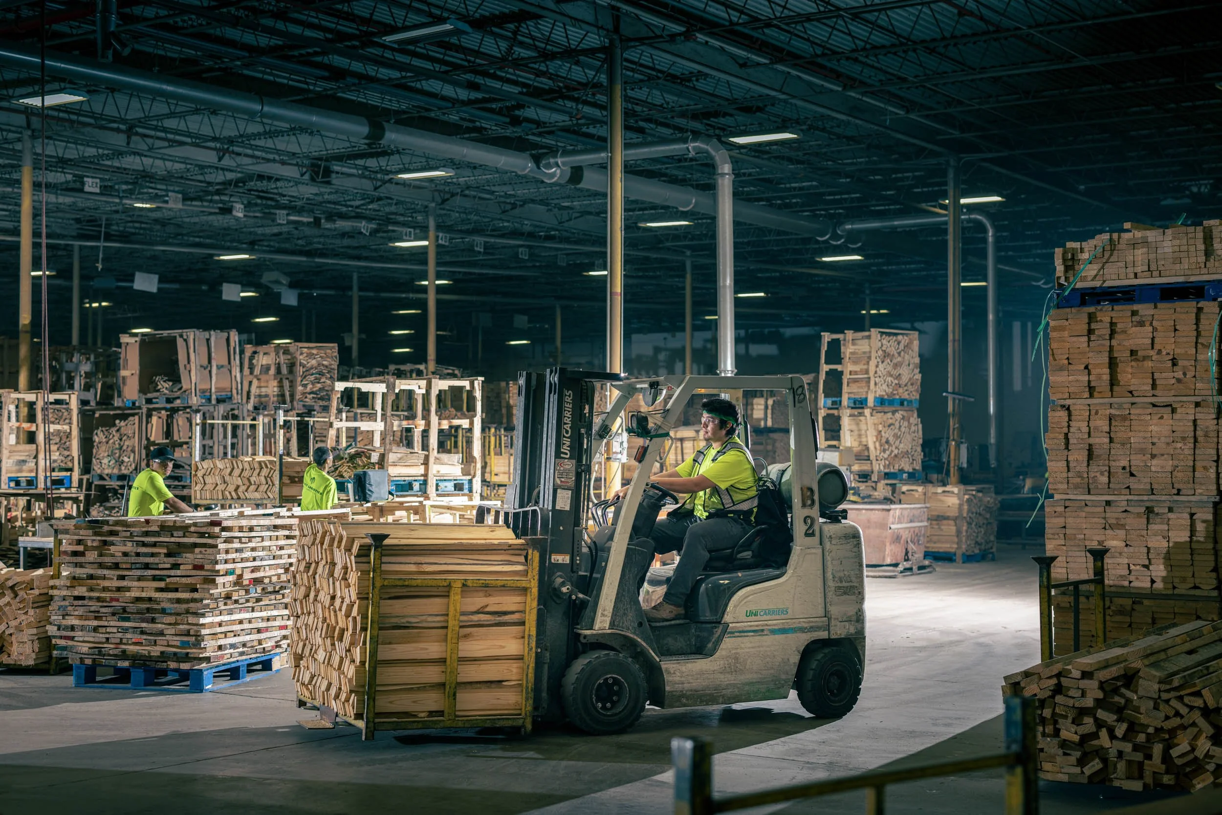 Workers operating forklifts and handling stacks of wooden planks in a large warehouse or industrial storage facility.