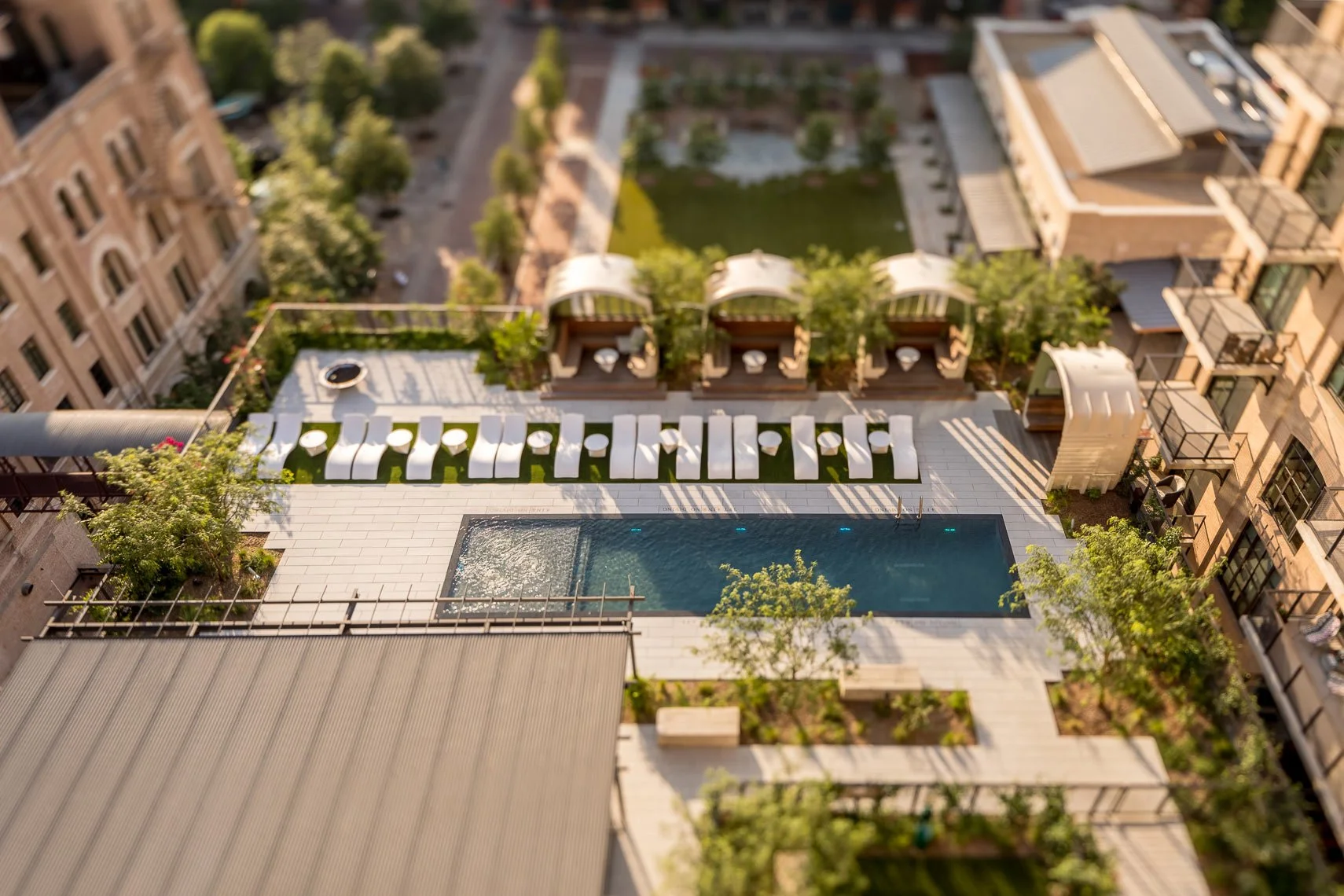 An aerial view of a modern residential rooftop with a swimming pool, lounge chairs, and shaded cabanas, surrounded by trees and other buildings.