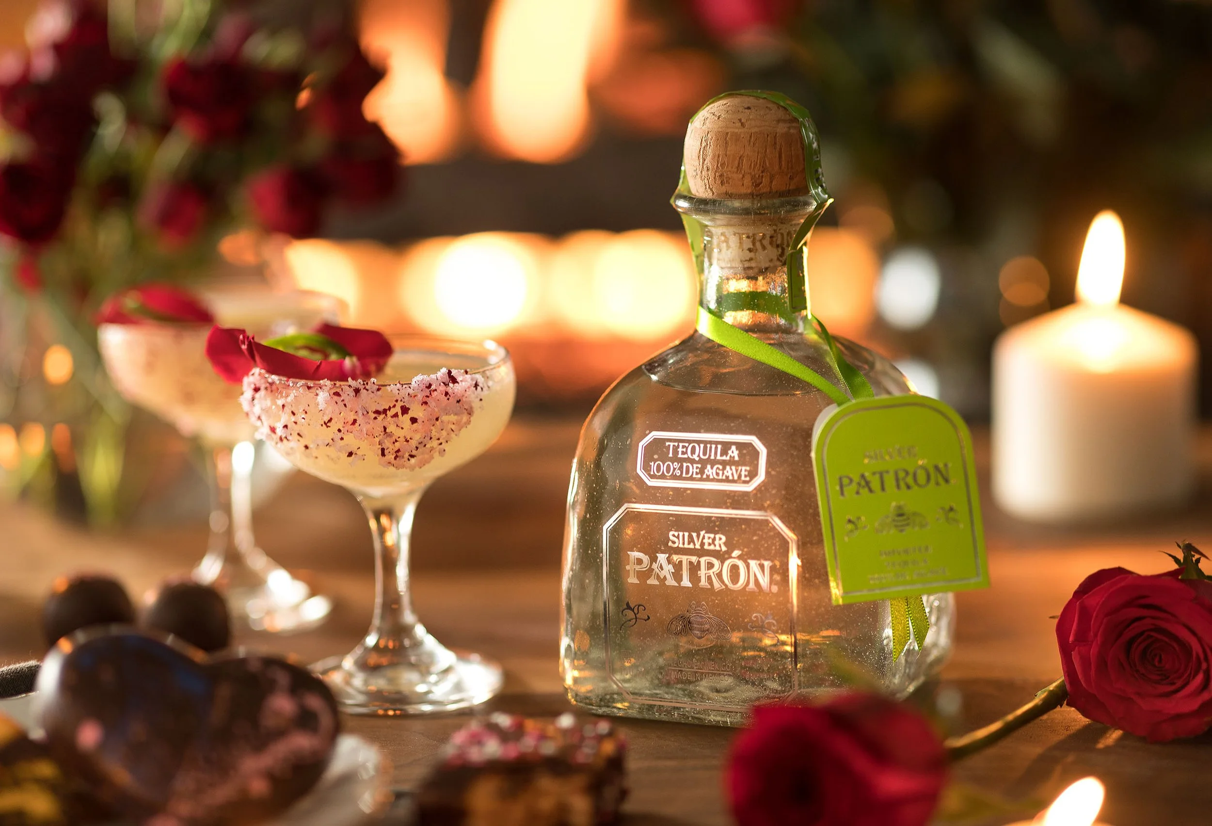 A bottle of tequila labeled 'Silver Patrón' is set on a wooden surface, surrounded by lit candles, roses, and small dark berries, creating an elegant and warm ambiance.