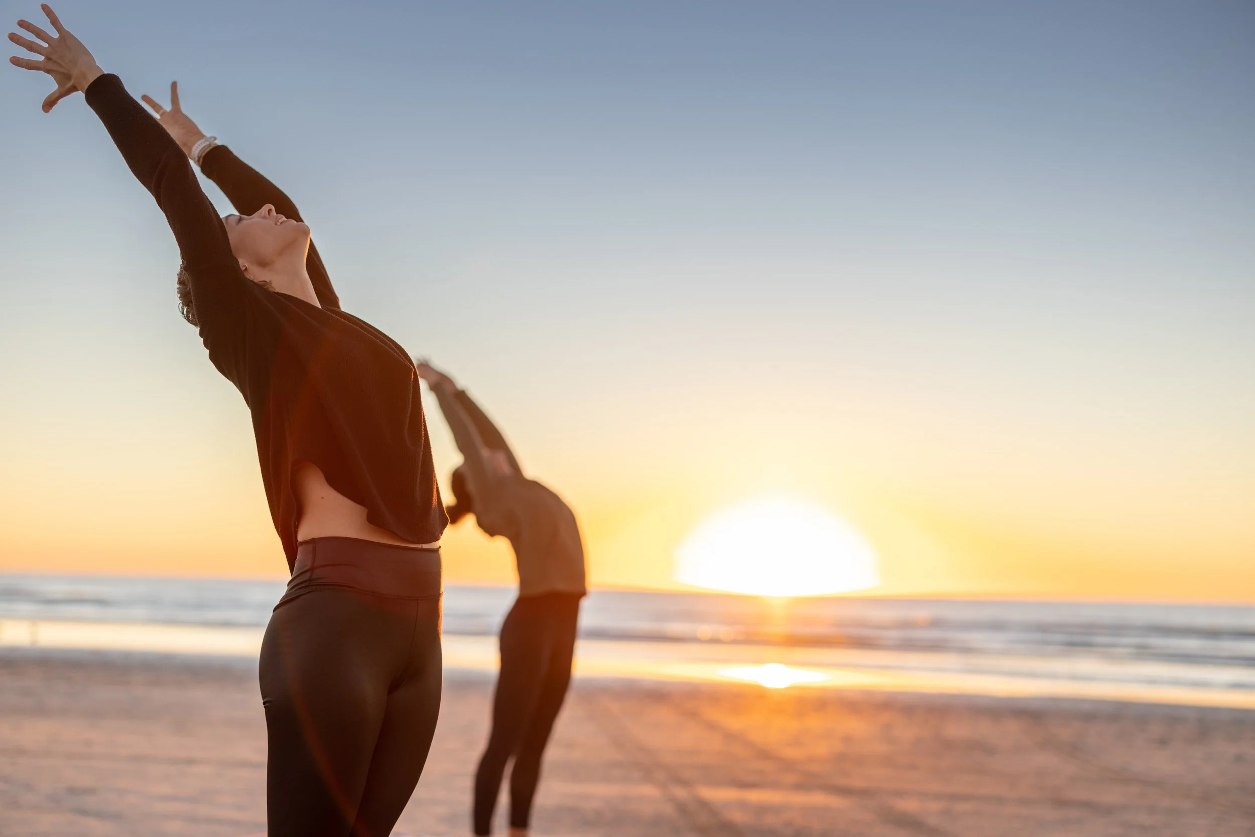 A person practicing yoga or meditation outdoors at sunset on the beach, with their arms raised and palms open, facing the sky.