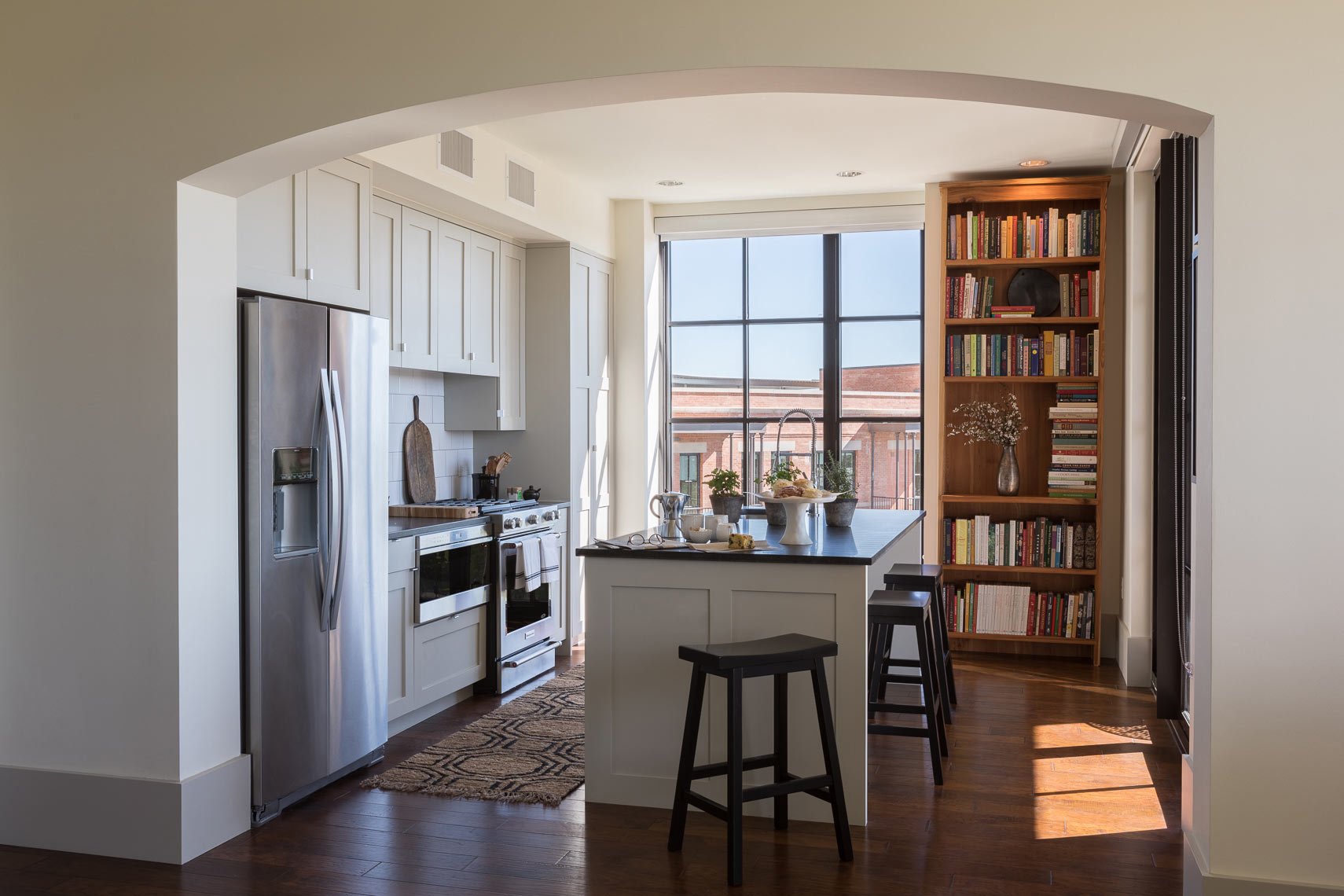 Modern kitchen with white cabinetry, stainless steel refrigerator, and a black countertop island with three stools. Large window behind the island, and a bookshelf with decorative items and plants on the right.