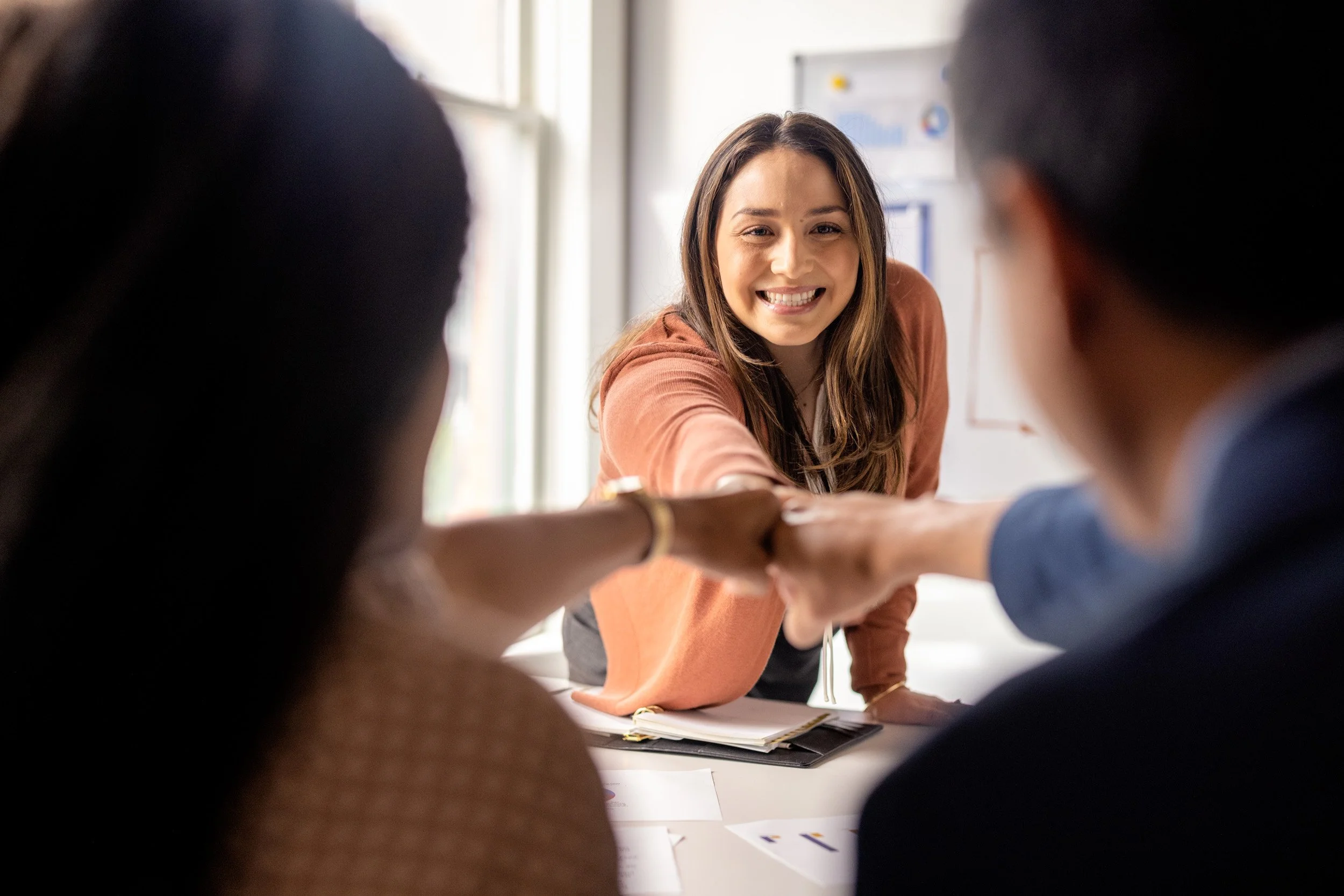A woman smiling and reaching out her hand in a group huddle with four other people around a table, in an indoor setting.