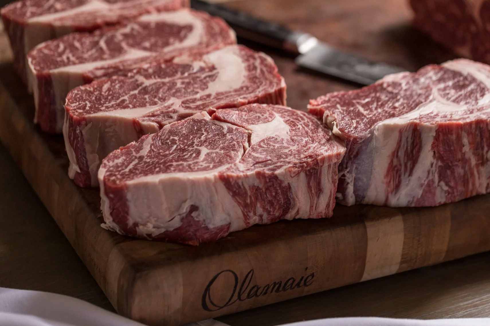 Six raw beef tomahawk steaks with marbling on a wooden cutting board.