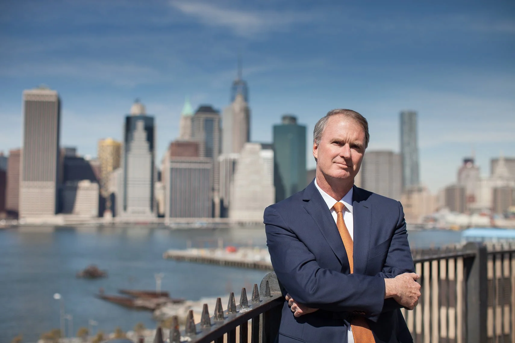 A middle-aged man in a navy blue suit with an orange tie stands with arms crossed in front of a city skyline from a waterfront perspective.
