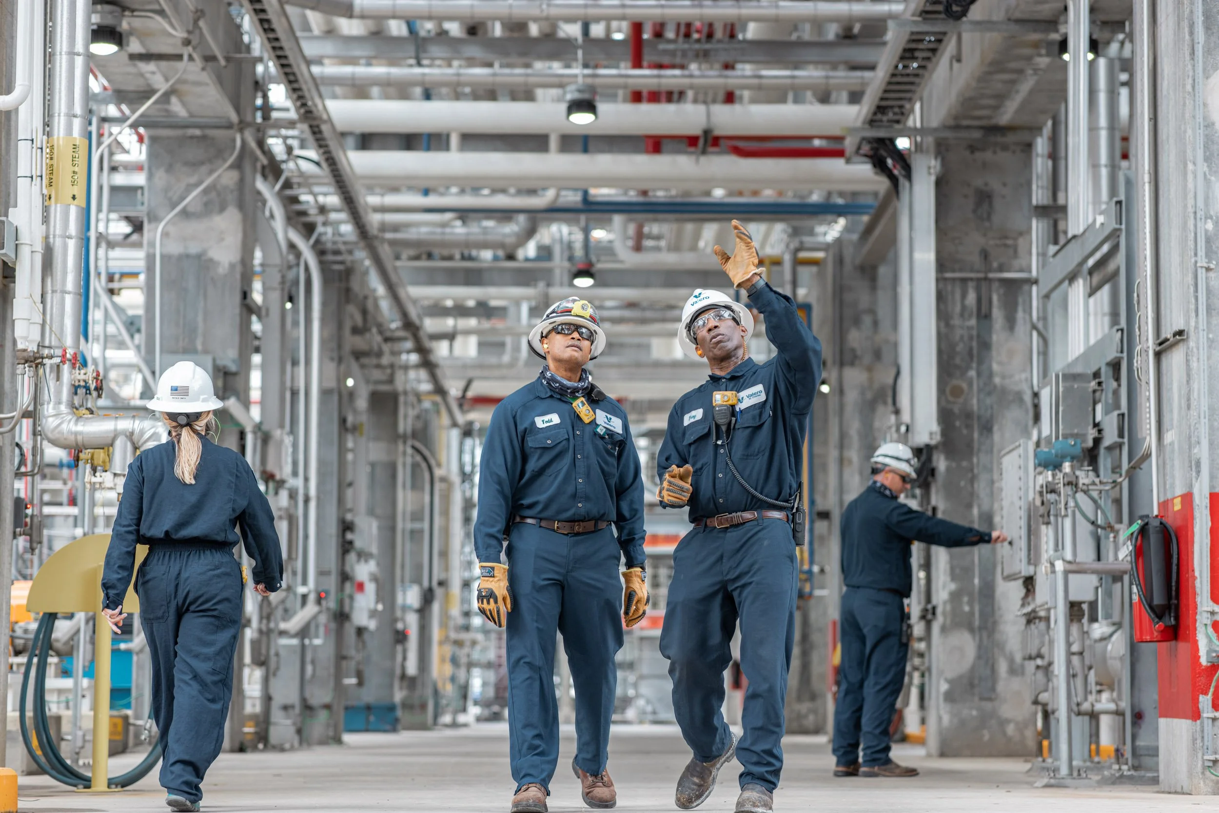 Four construction workers wearing helmets and safety gear inspecting a large industrial facility with exposed pipes and construction materials.