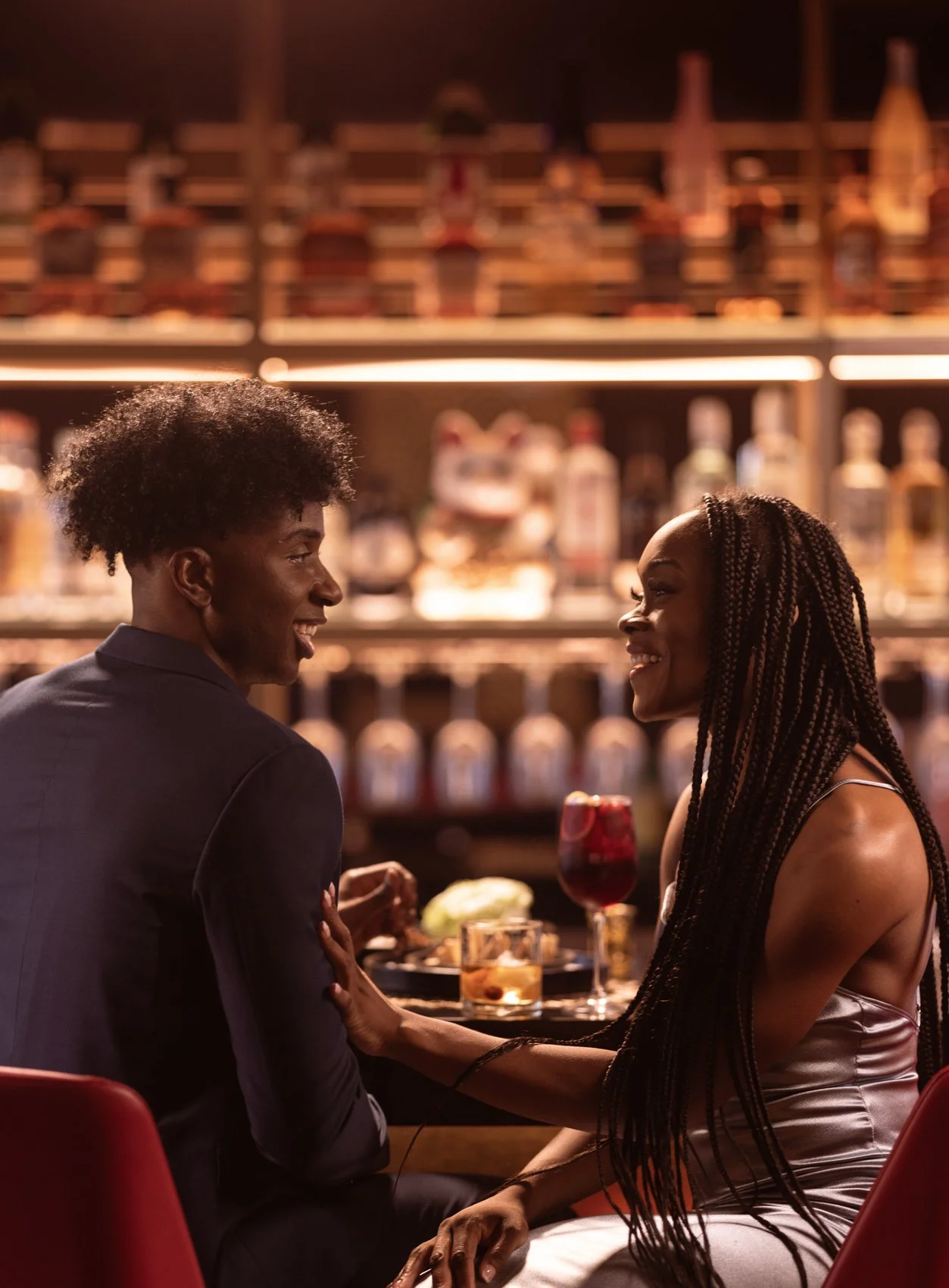 A man and woman are sitting at a bar, facing each other and smiling. The woman is touching the man's arm and they are holding hands. There are drinks on the table, and bottles of alcohol can be seen on shelves behind them.