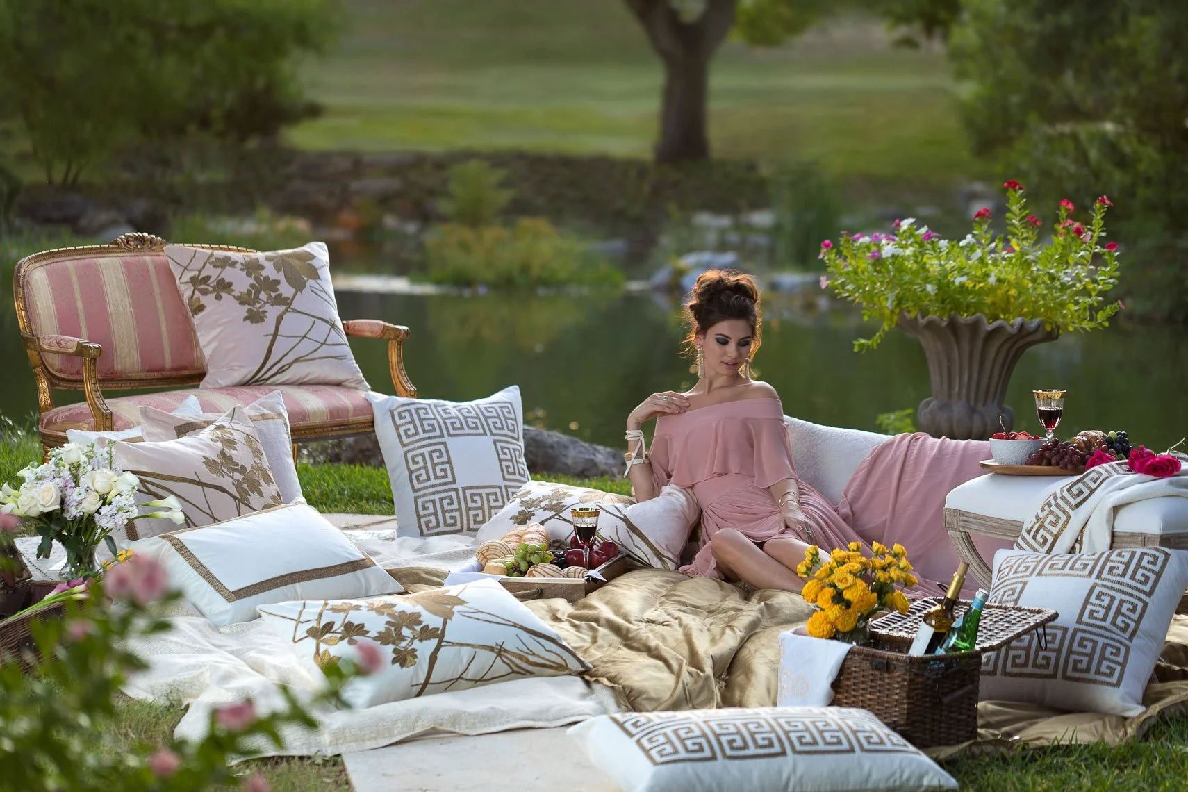 A woman in a pink dress sits on a mattress surrounded by pillows and flowers outdoors near a pond, with a tray of food and drinks, and a large decorative flowerpot with pink flowers nearby.