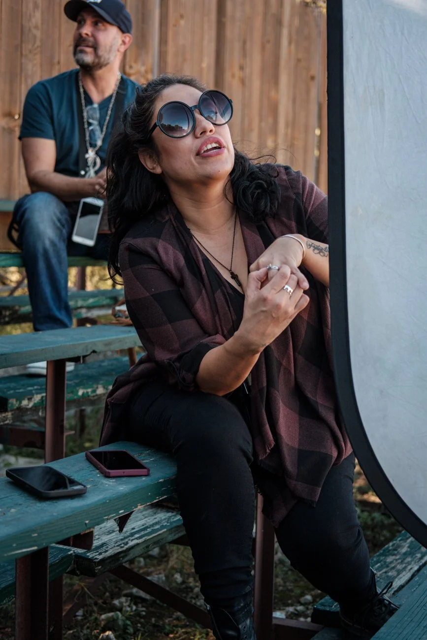 A woman with dark hair, wearing sunglasses and a plaid shirt, is sitting outdoors on a bench, looking at a large reflector. A man with a beard and wearing a navy shirt and cap is sitting behind her, looking away. There are two smartphones on the benc