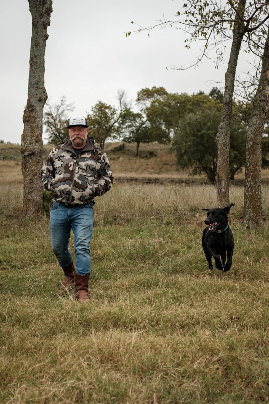 A man walking with a black dog in an open grassy area with trees and overcast sky.