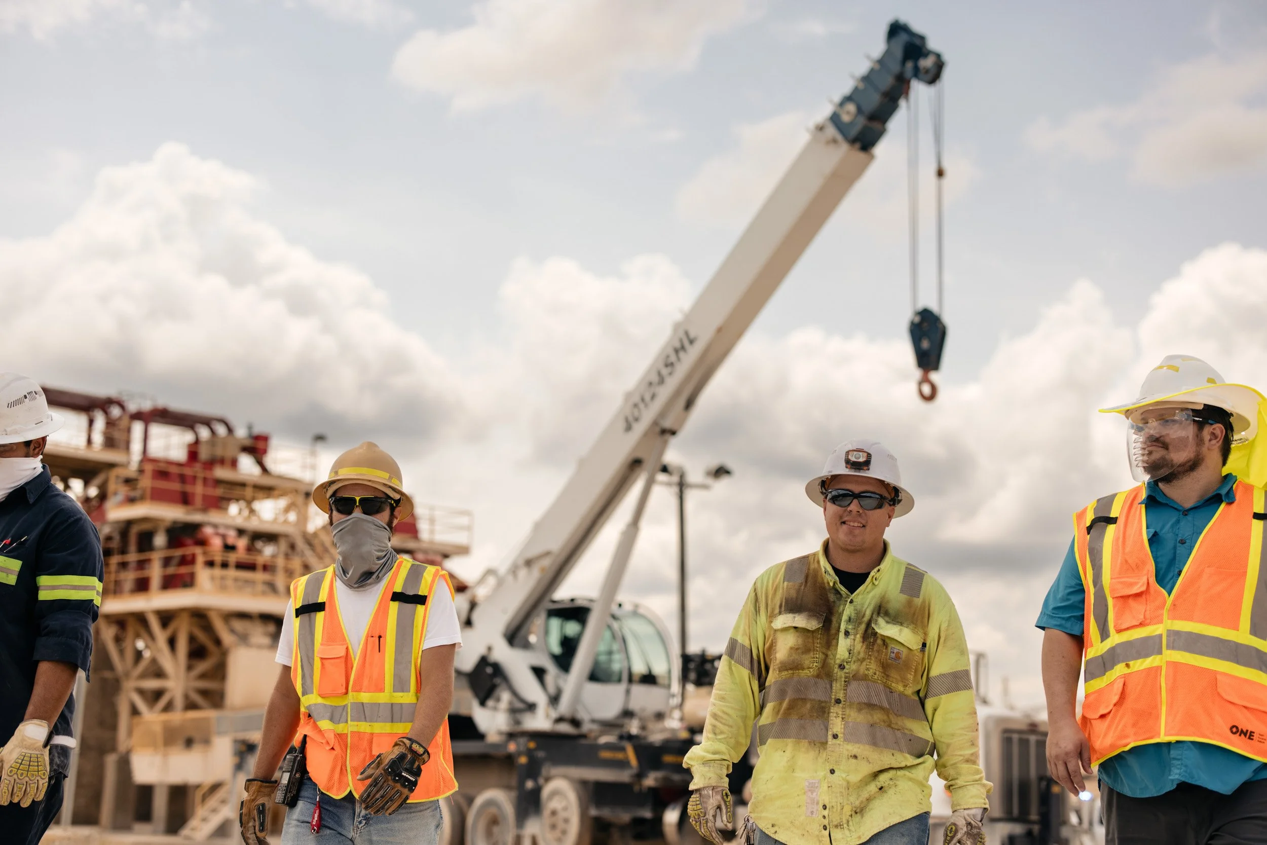 Construction workers wearing safety vests, helmets, and gloves standing outdoors with a crane and building framework in the background under a partly cloudy sky.