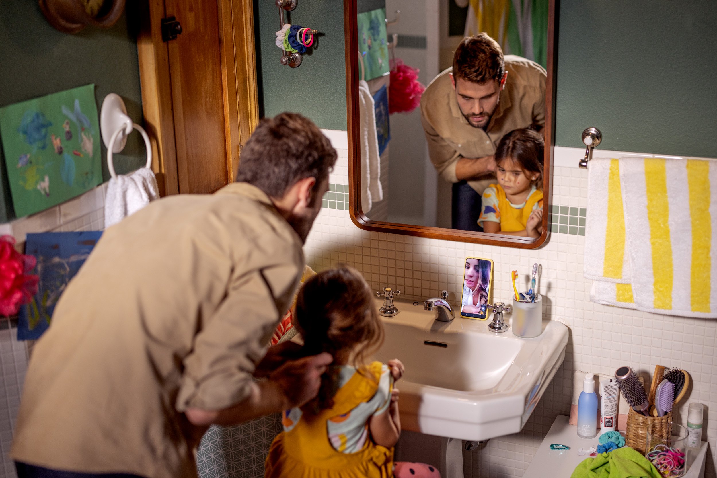 A man watches a woman and two children in a bathroom mirror as they wash their faces. The bathroom has a green wall, a white sink, and various toiletries and colorful towels.