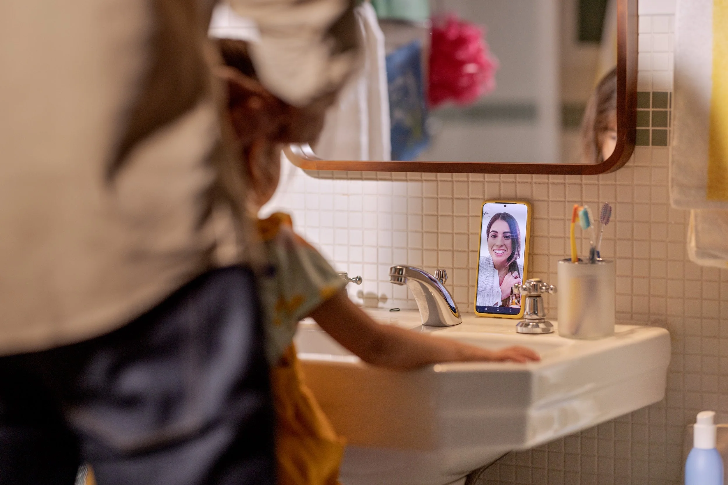 A woman is washing her hands at a bathroom sink, with a smartphone showing her smiling face placed on the counter in front of her.