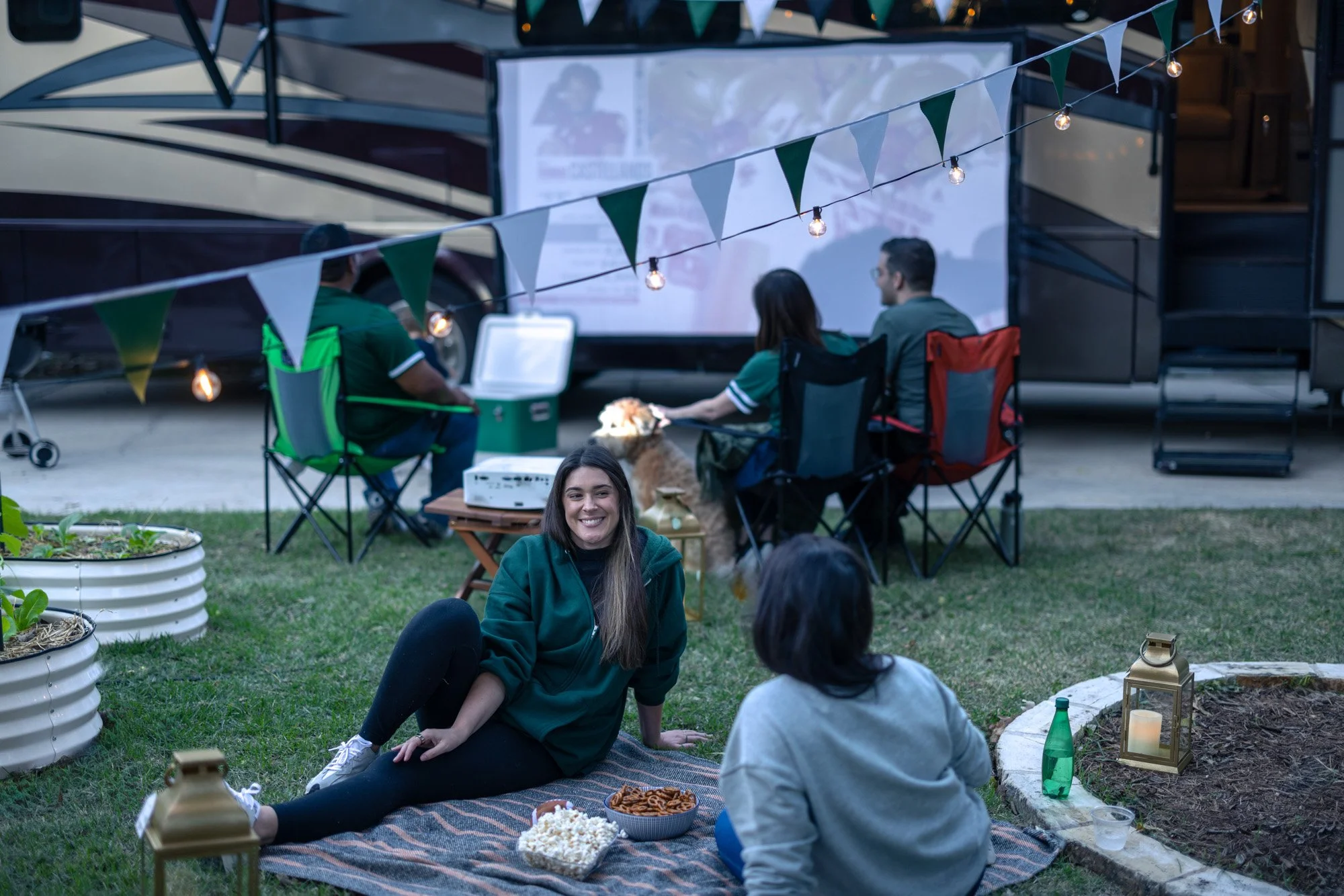 A group of people enjoying a gathering outdoors in a backyard during evening. Some are seated on chairs and a blanket on the grass, with food and drinks on the ground, while others sit around a picnic table. String lights and bunting add to the festi