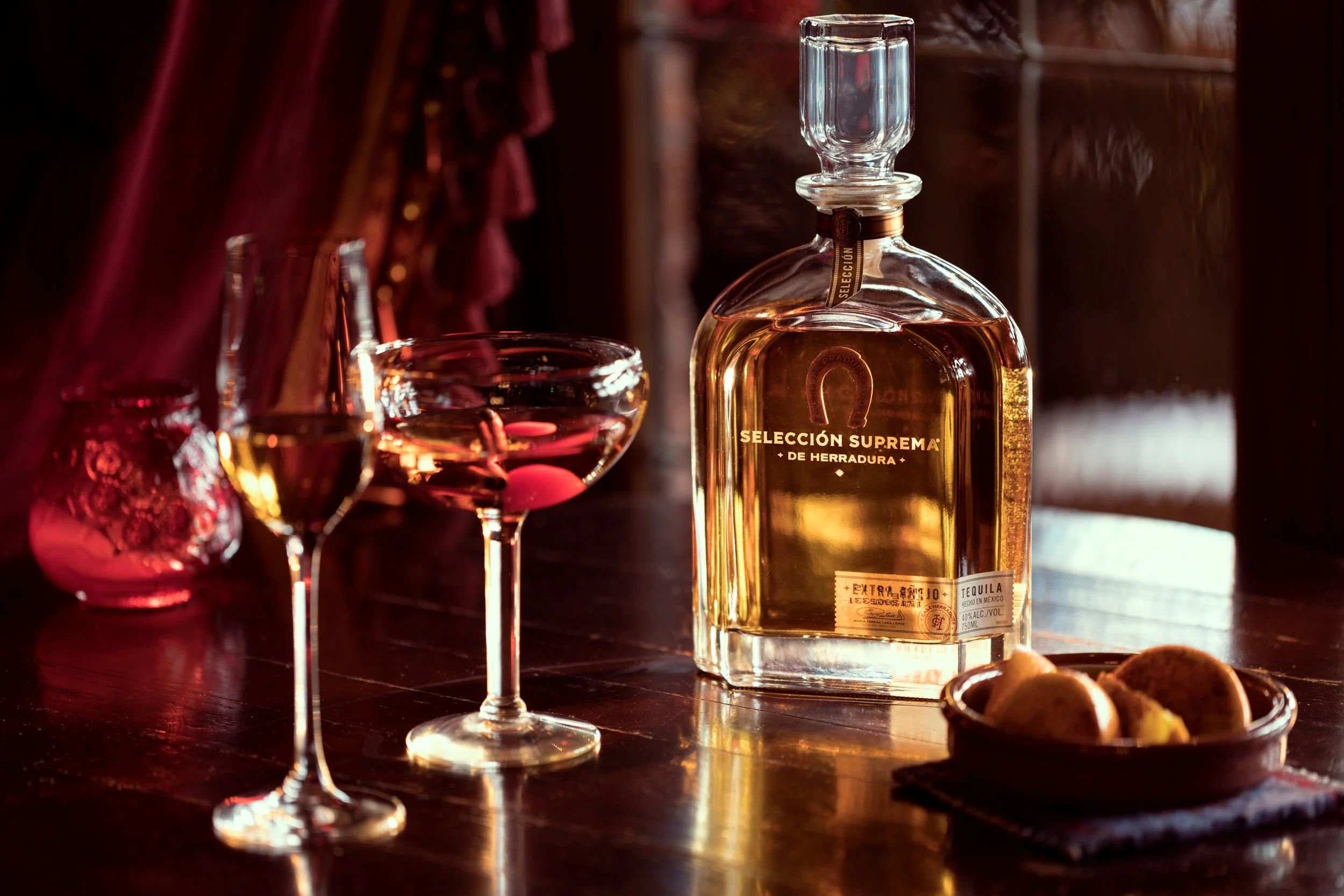 A bottle of tequila and two glasses with pink-colored drinks on a dark wooden surface, with a plate of nuts nearby and a dark, rich background.