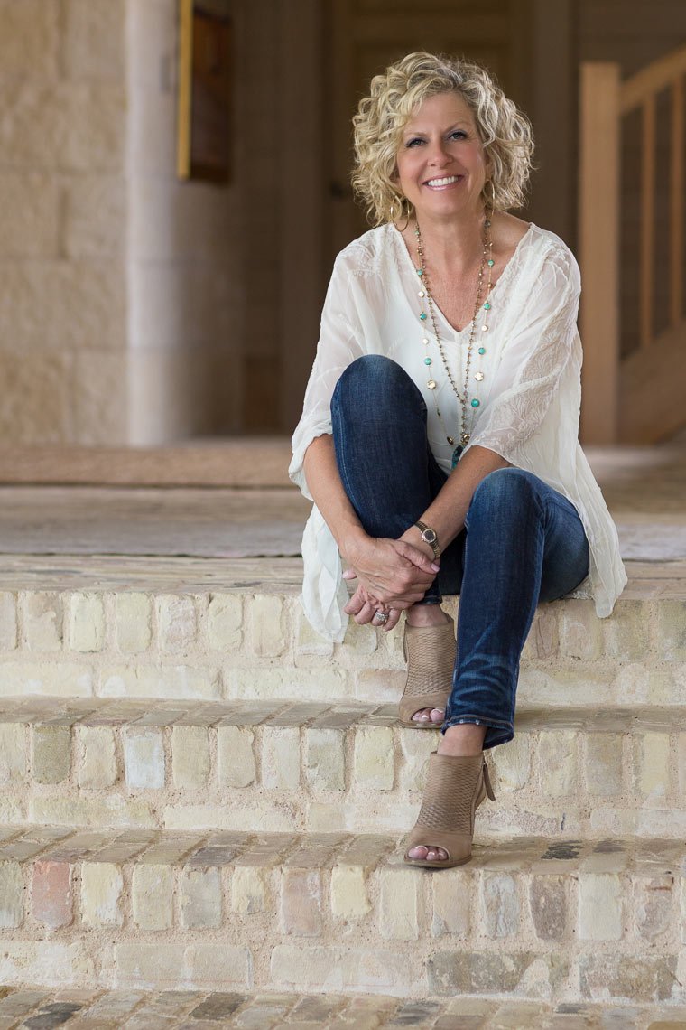 A woman with curly blonde hair sitting on brick stairs, smiling, wearing a white blouse, blue jeans, taupe open-toe ankle boots, and layered necklaces, in a rustic indoor setting.