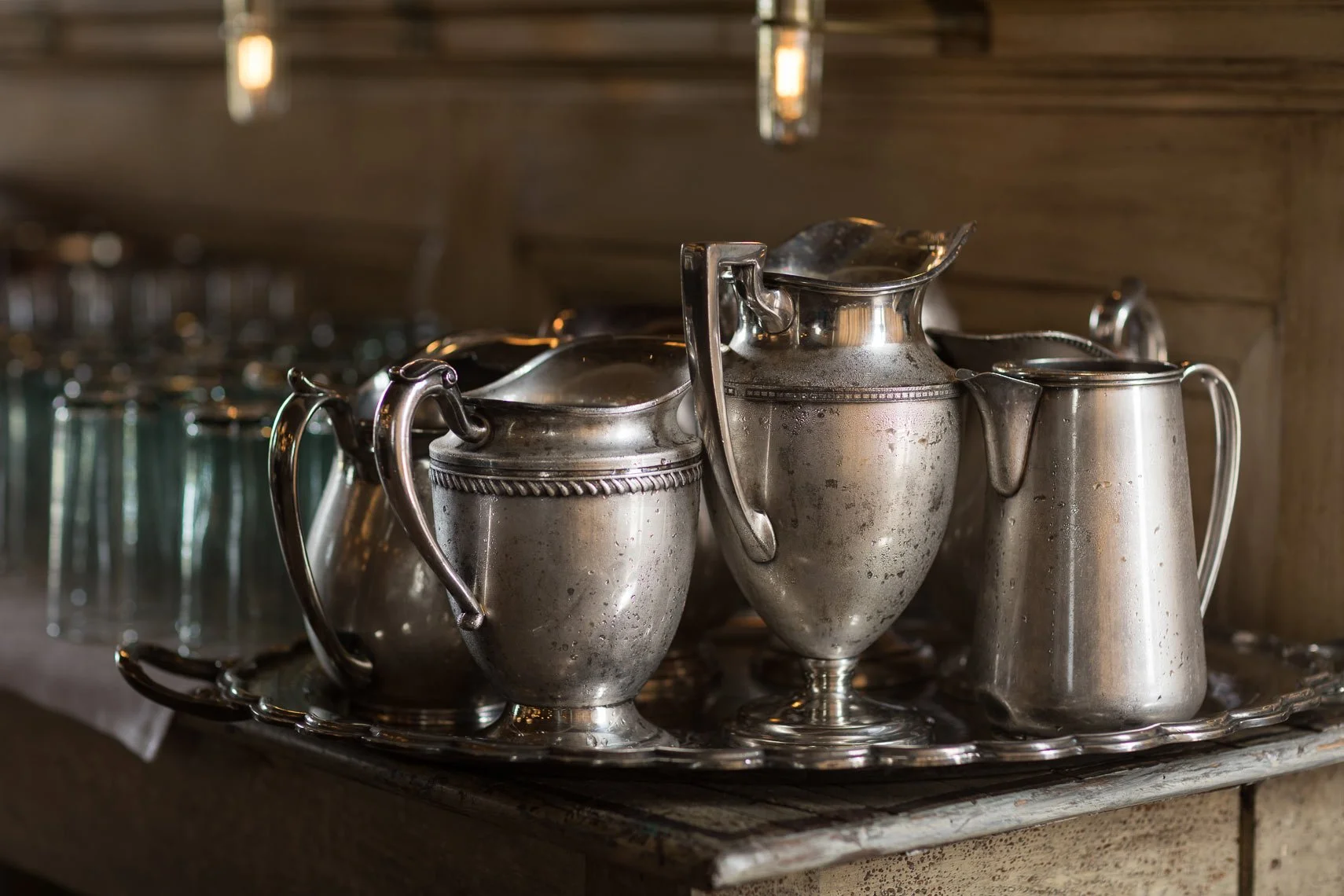 A silver tray holding four vintage silver pitchers of varying sizes, with the middle one featuring a decorative handle, set against a wooden background.