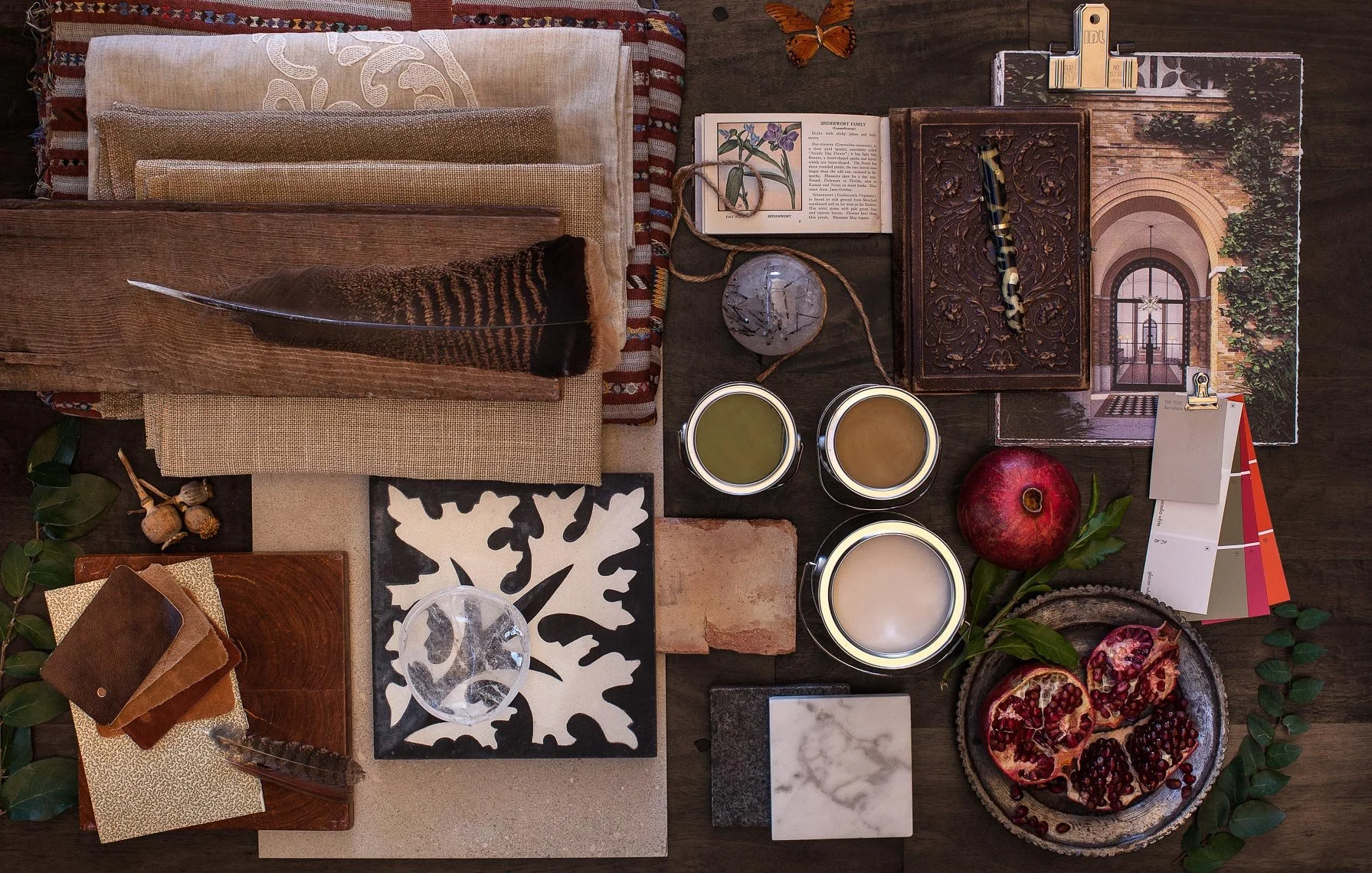 A flat lay of interior design and decor items including fabric swatches, a feather, books, jars, a pomegranate, tiles, a decorative box, paint swatches, a photograph, and various plants on a wooden surface.