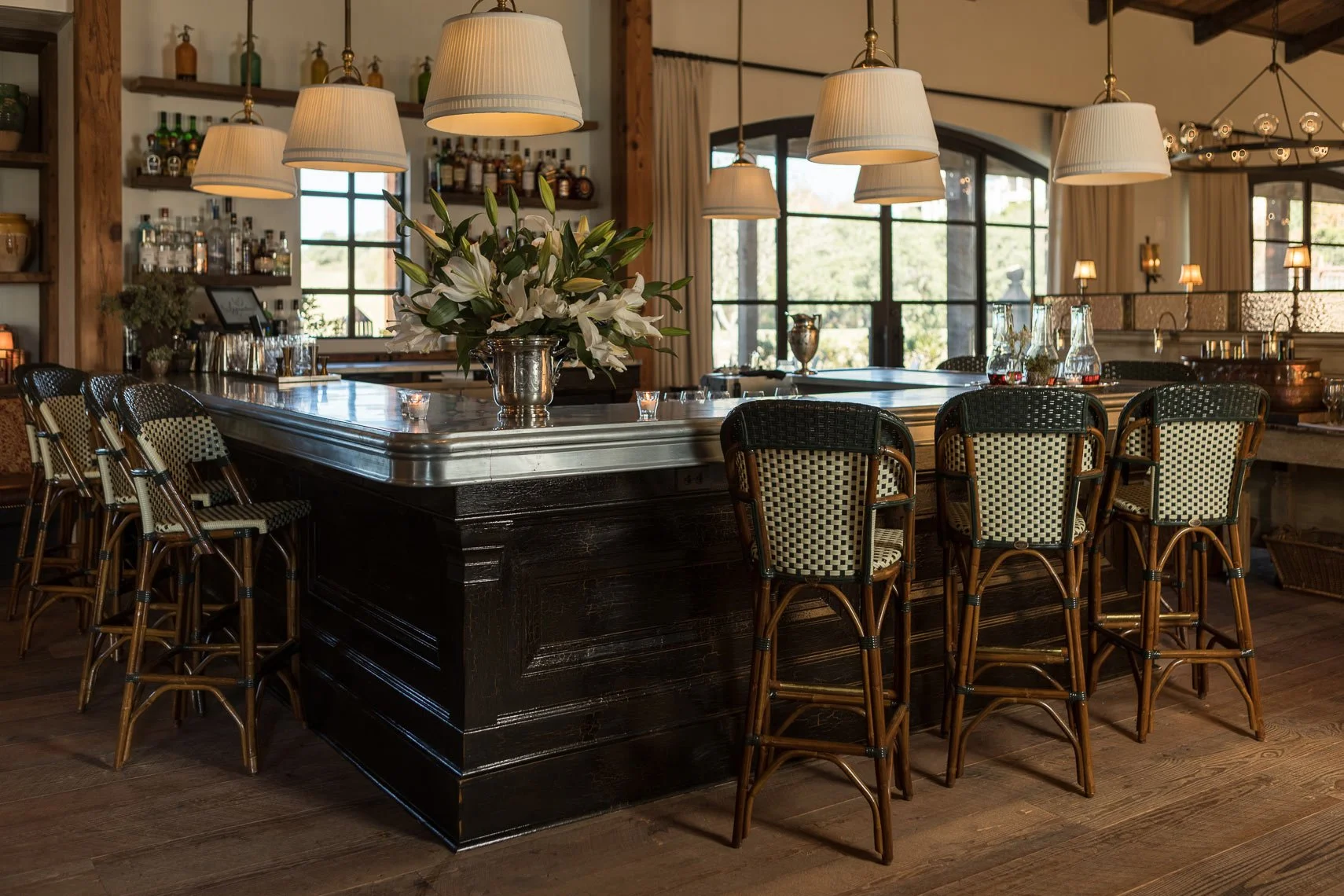 A cozy restaurant interior with a dark wooden bar counter, chairs with woven patterns, hanging lamps, and a large floral arrangement of white lilies in a silver vase.