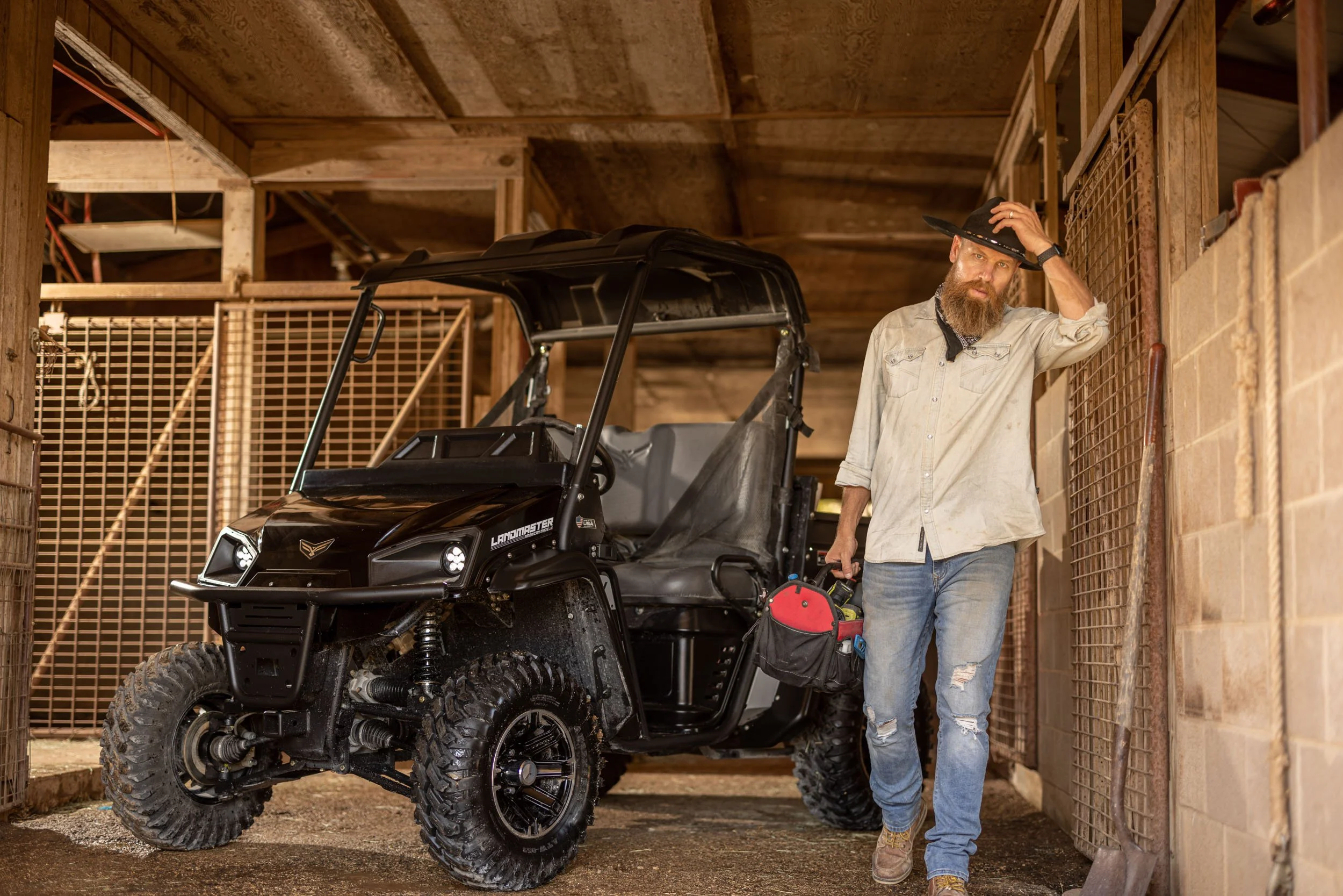 Man with a beard wearing a cowboy hat, beige shirt, and ripped jeans standing next to a black off-road utility vehicle inside a wooden barn or shed, holding a tool in his right hand and touching his hat with his left hand.