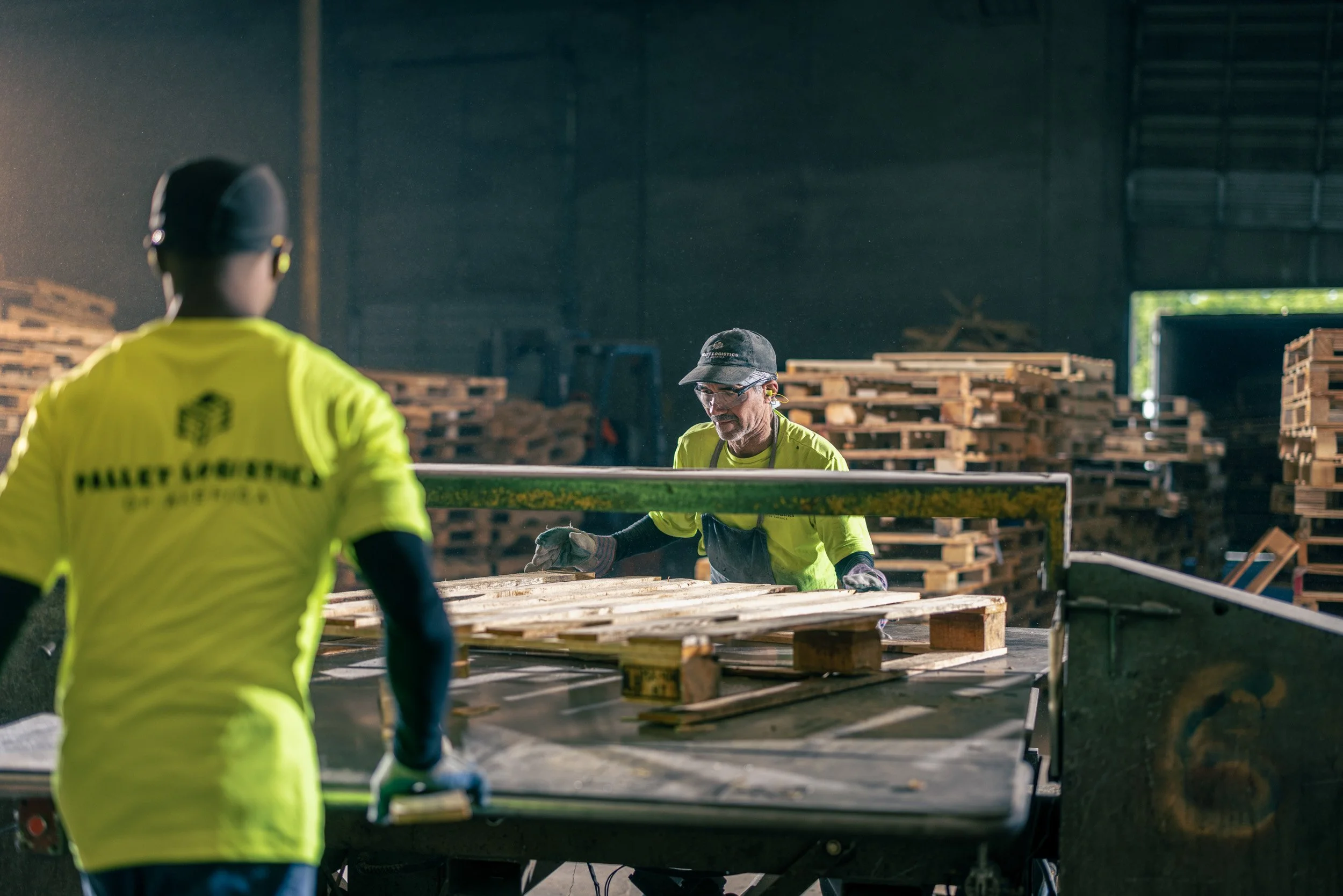 Workers in yellow safety shirts inside a warehouse stacking wooden pallets, with sunlight shining through an open door in the background.