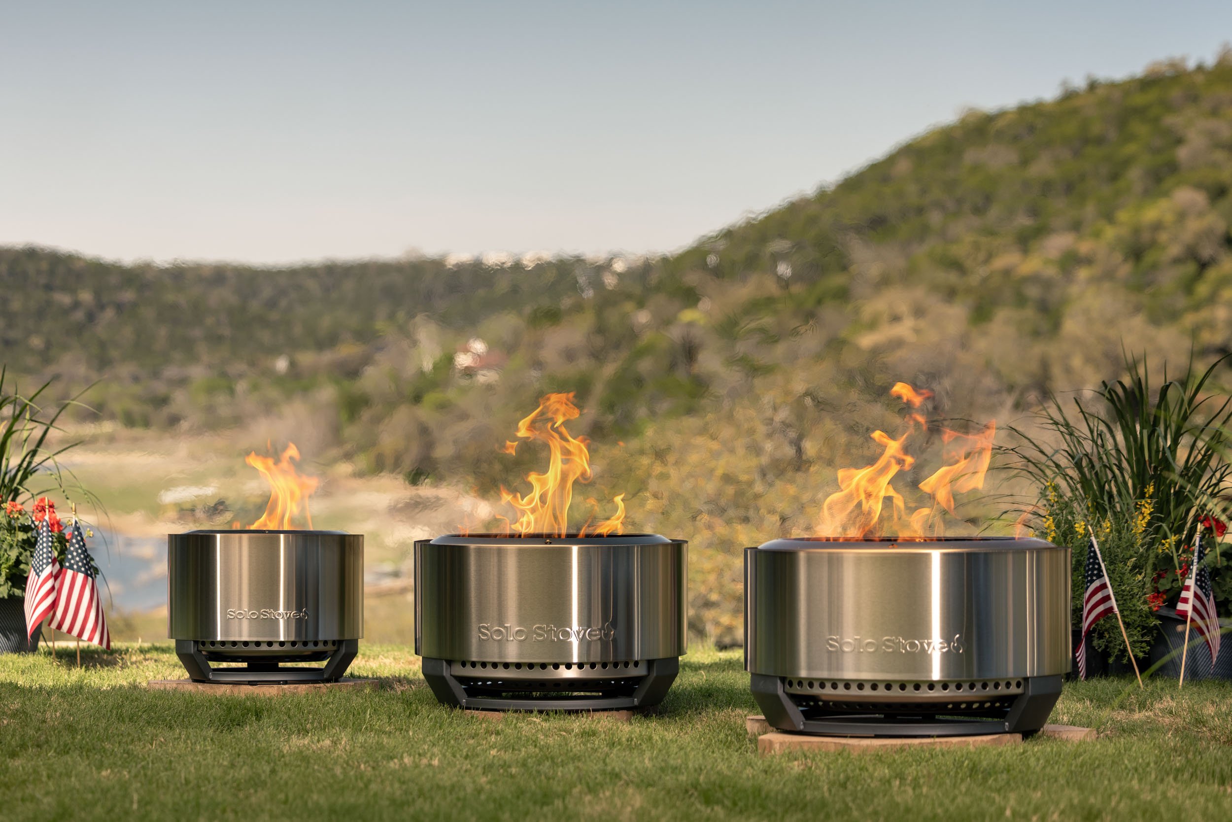 Three stainless steel Solo Stove fire pits outdoors on a grassy area with a scenic hillside background, lit with flames, surrounded by small American flags and patriotic decorations.