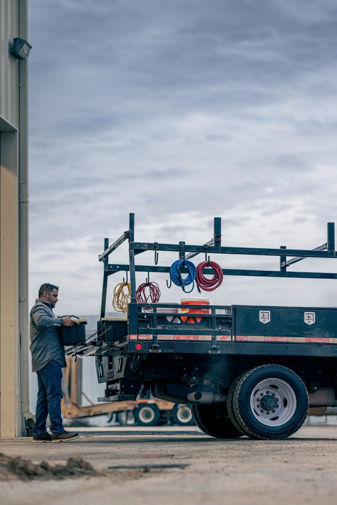A man standing next to a utility truck with hoses and equipment, holding a toolbox, under a cloudy sky.