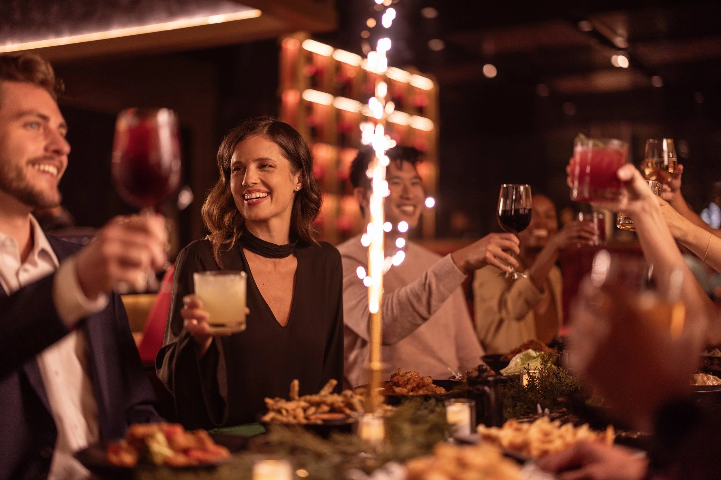People celebrating at a dinner party with drinks and food, with sparklers in the background.