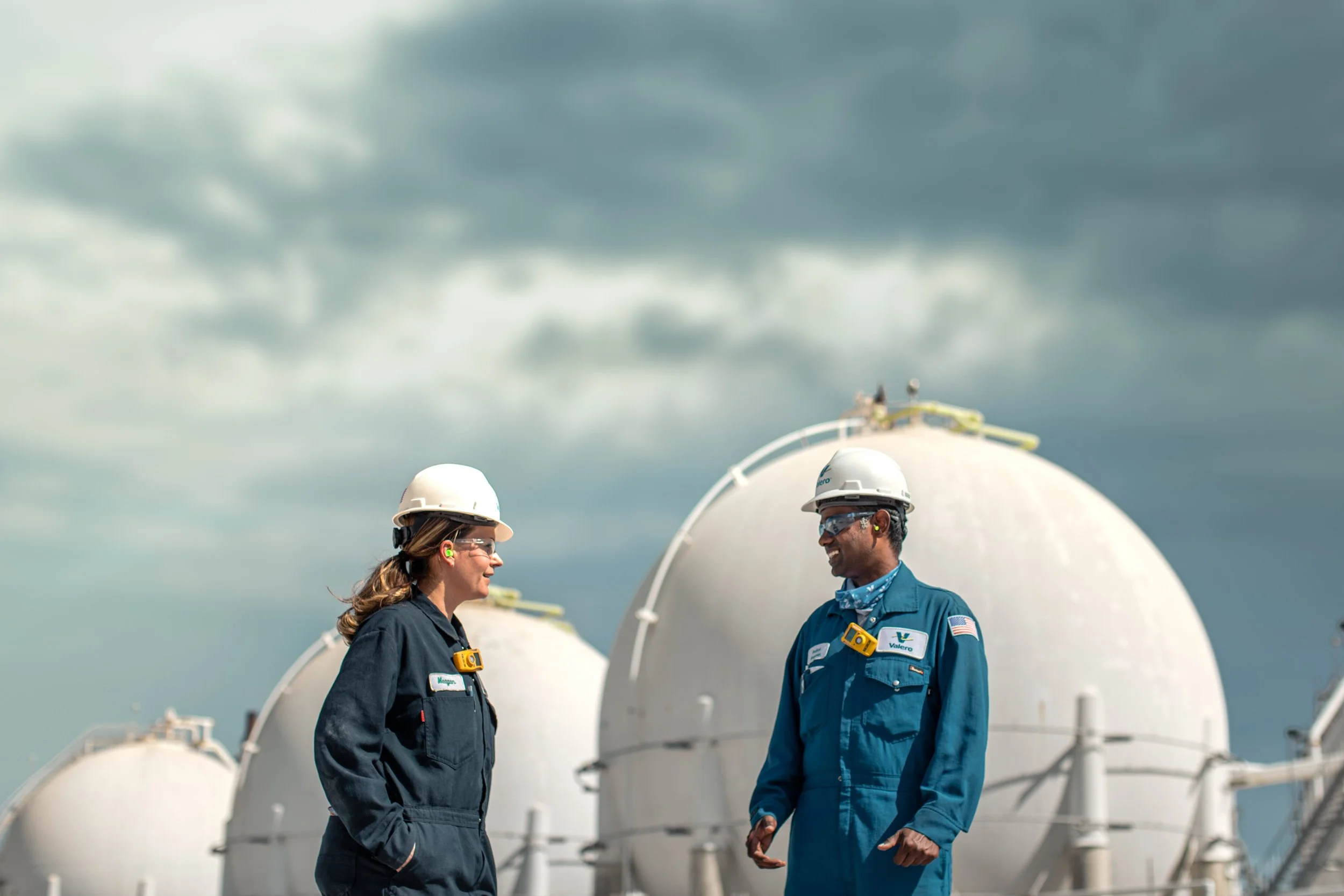 Two workers in safety gear and hard hats conversing outdoors near large white industrial tanks under a cloudy sky.