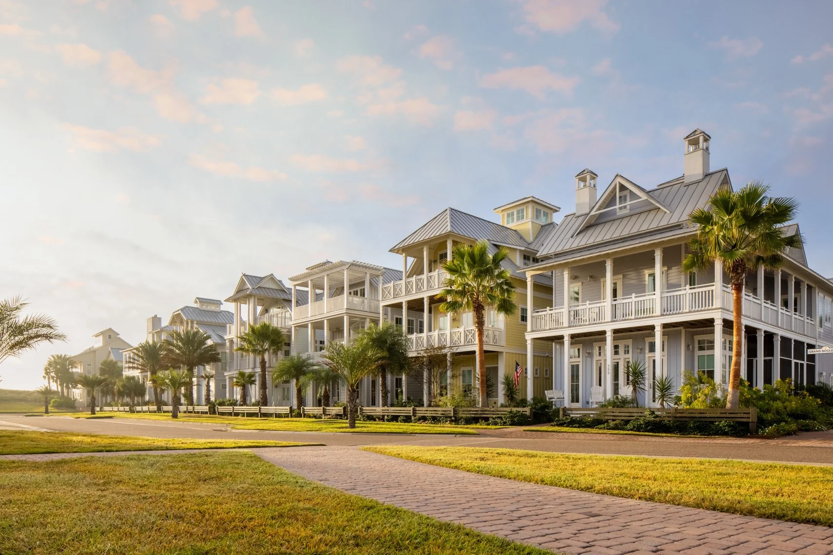 A row of multi-story beach houses with porches, palm trees lining the street, and a blue sky with soft clouds.
