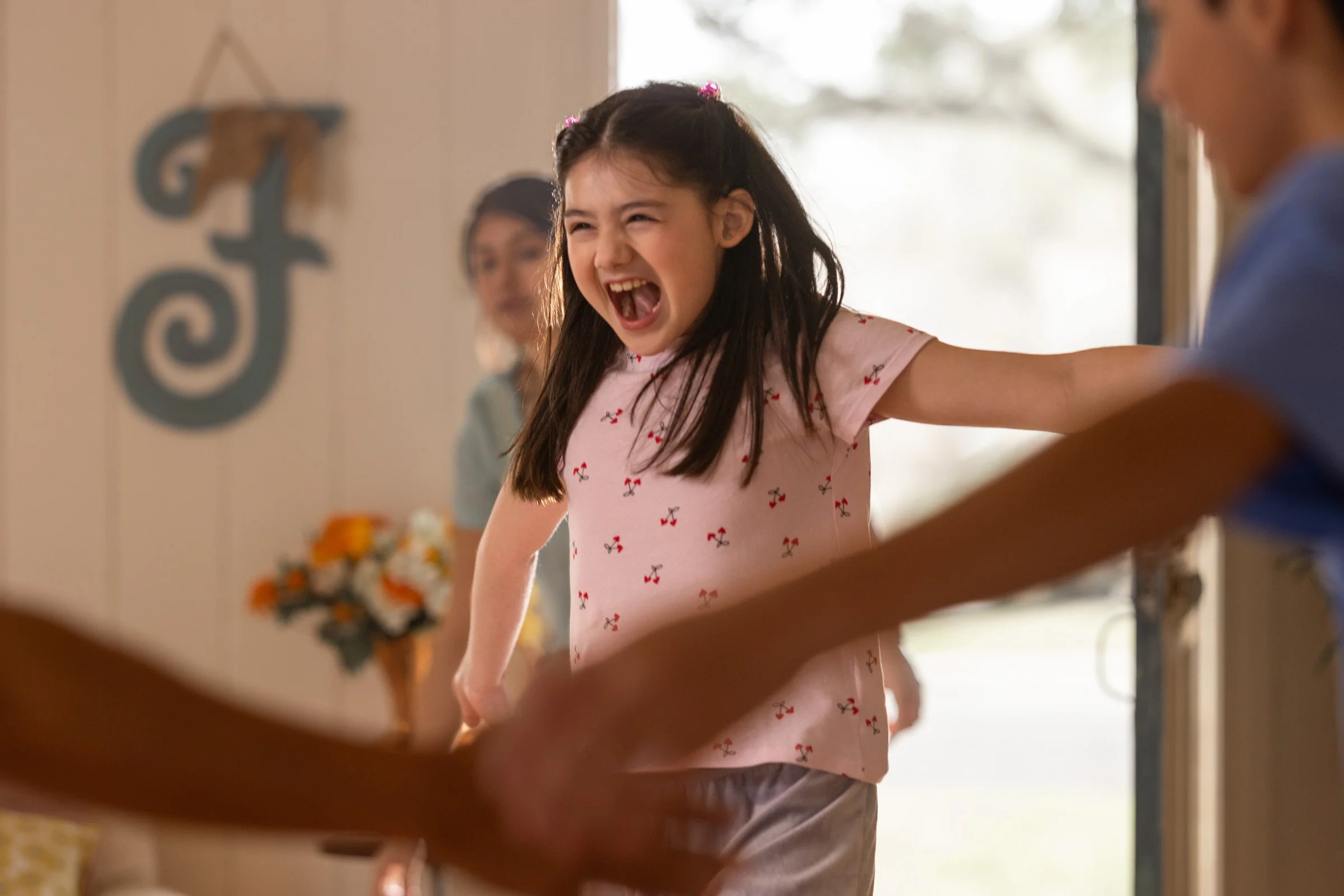 A young girl with dark hair and wearing a pink shirt with cherry patterns, smiling and opening her mouth wide, with other children visible in the background and a decorative element with a miniature horse on the wall.